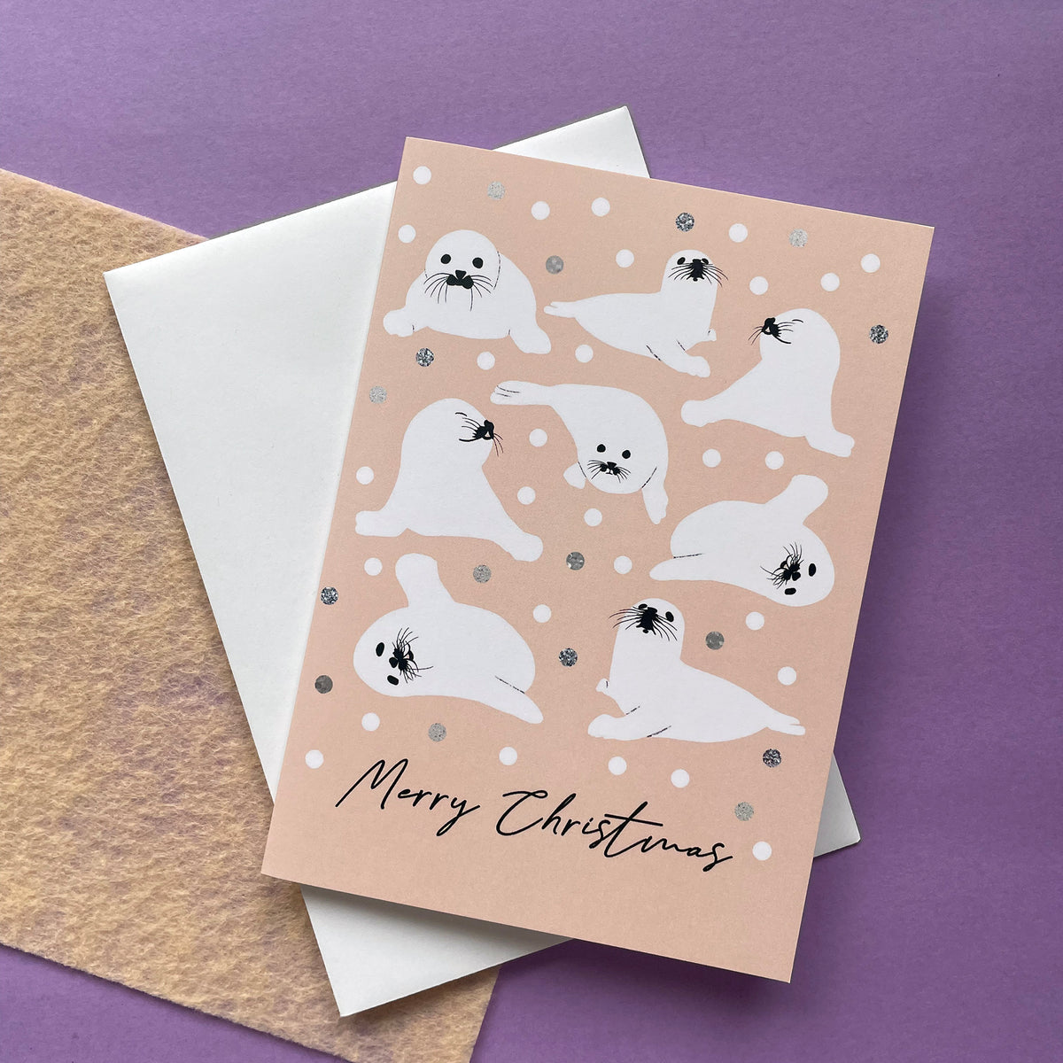 A Christmas greeting card featuring cute white seal illustrations on a peach background with snowballs, layered over a white envelope and a textured kraft paper sheet on a purple surface.