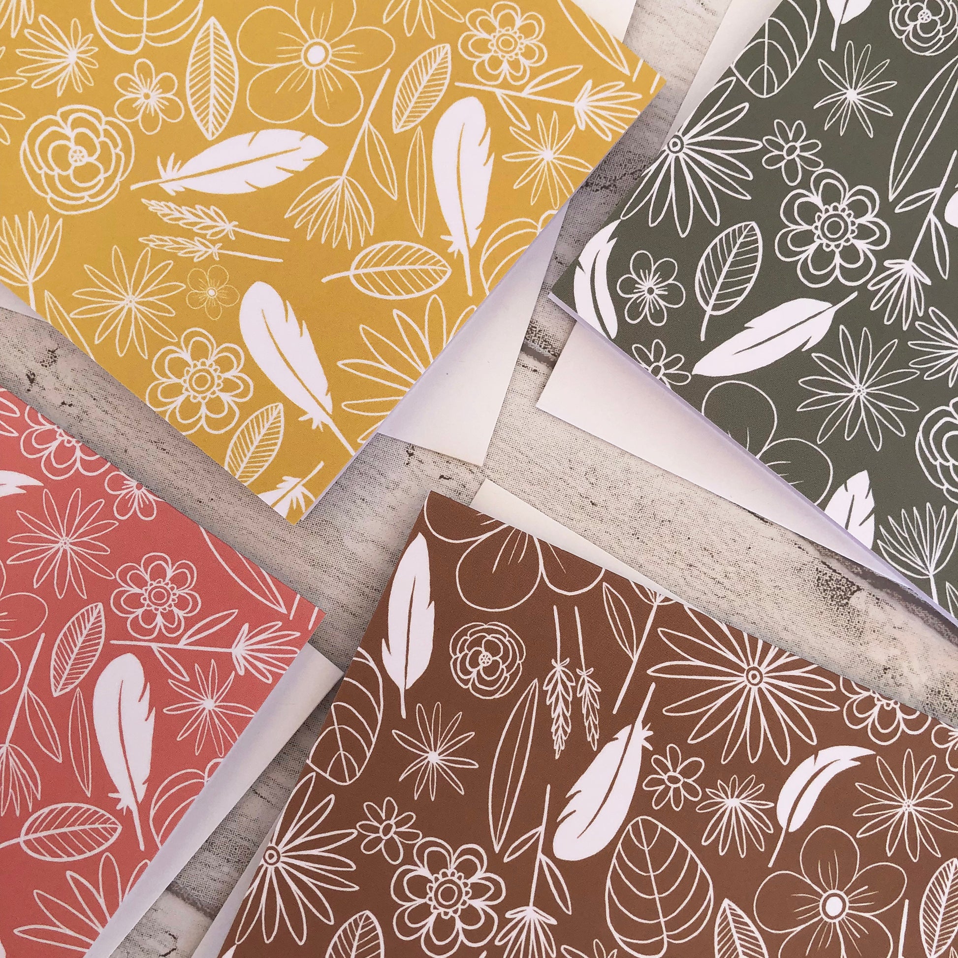 Image shows a collection of 4 notecards in pastel colours with white illustrations of flowers, feathers and leaves.