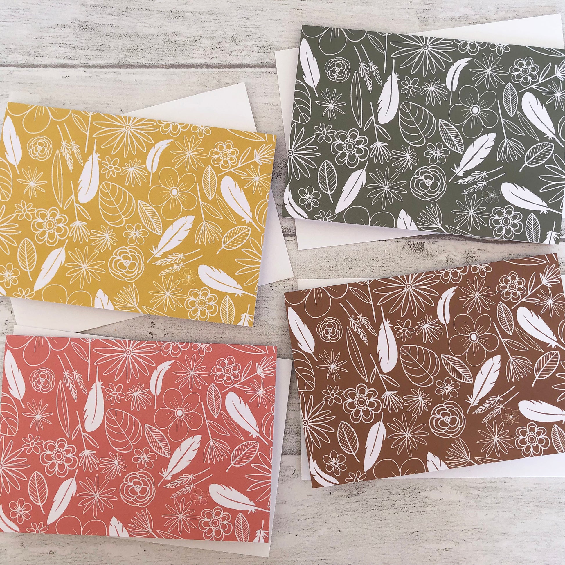 Image shows a collection of 4 notecards in pastel colours with white illustrations of flowers, feathers and leaves.