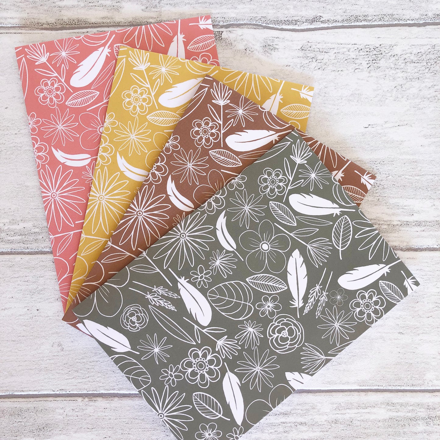 Image shows a collection of 4 notecards in pastel colours with white illustrations of flowers, feathers and leaves.