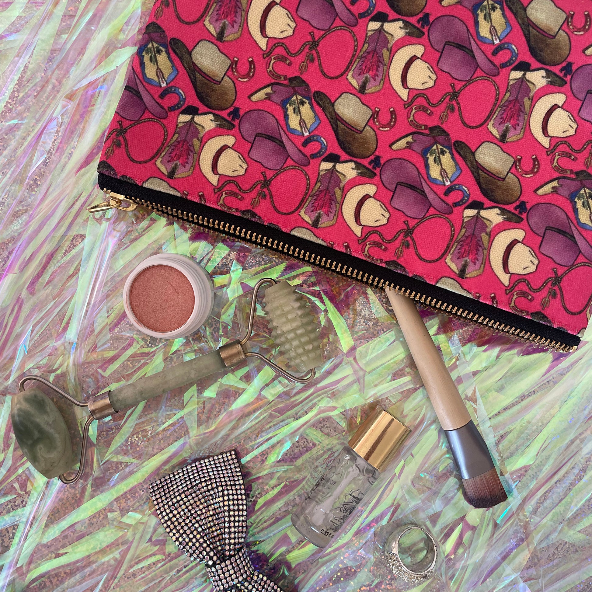 Image shows a bright cherry pink zip pouch with a fun western themed design, featuring cowgirl accessories, hats and boots.