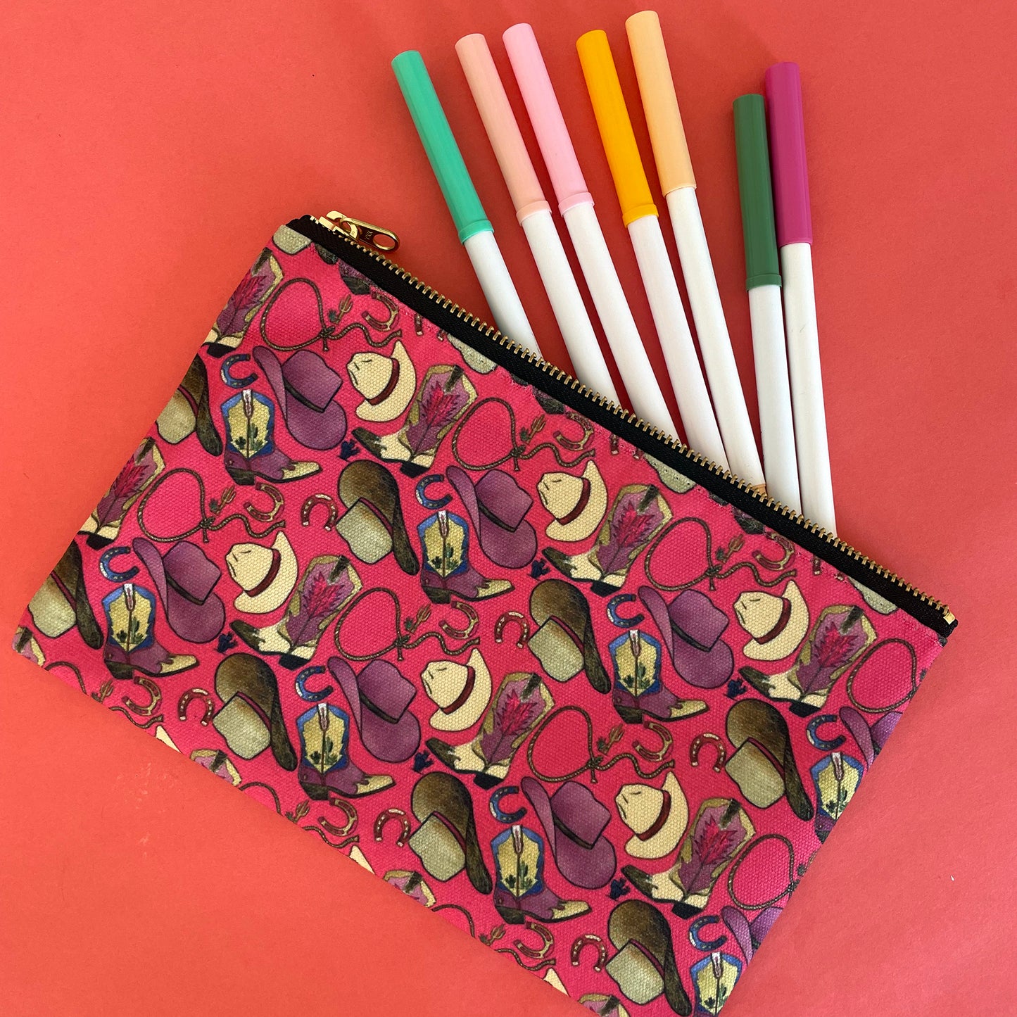 Image shows a bright cherry pink zip pouch with a fun western themed design, featuring cowgirl accessories, hats and boots.