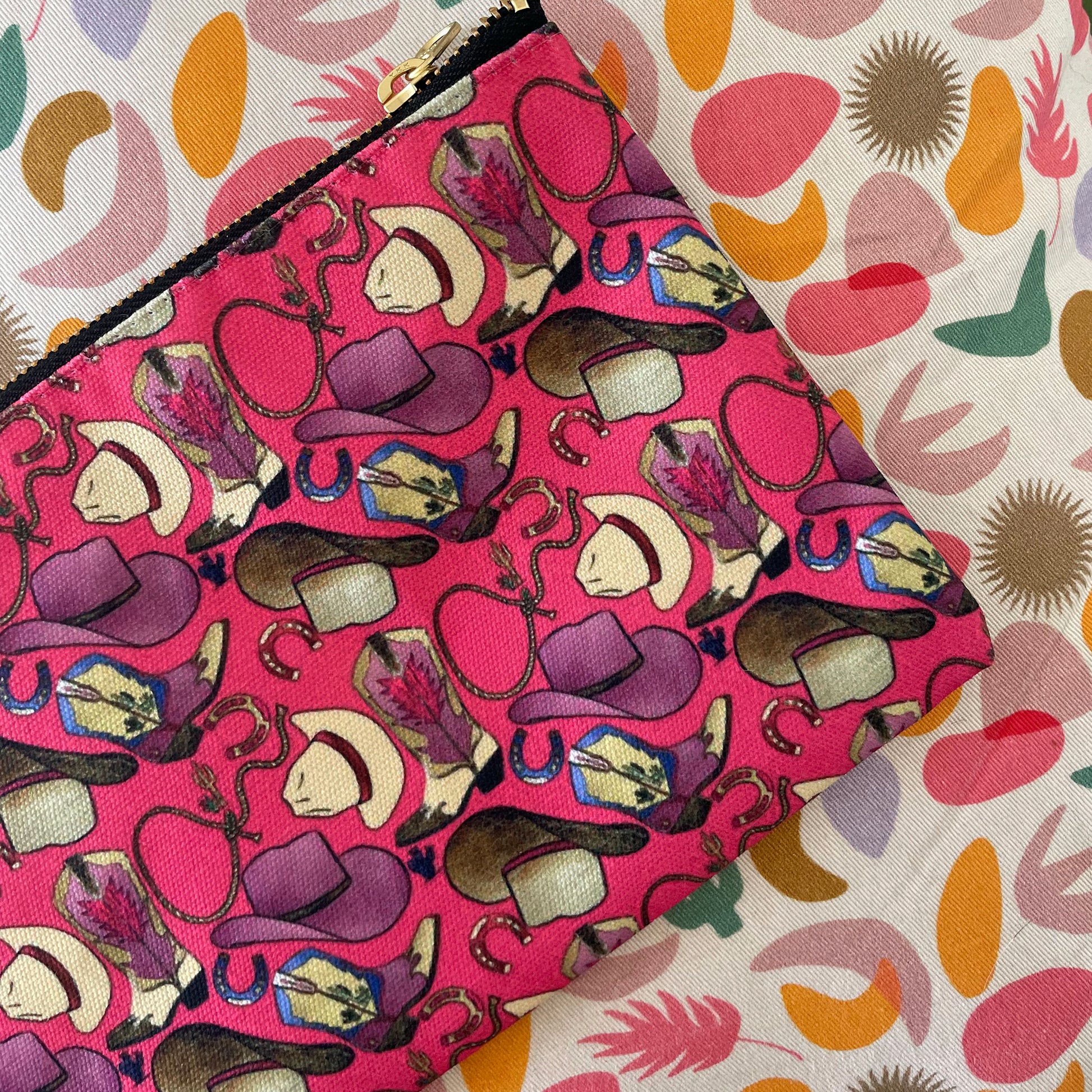 Image shows a bright cherry pink zip pouch with a fun western themed design, featuring cowgirl accessories, hats and boots.
