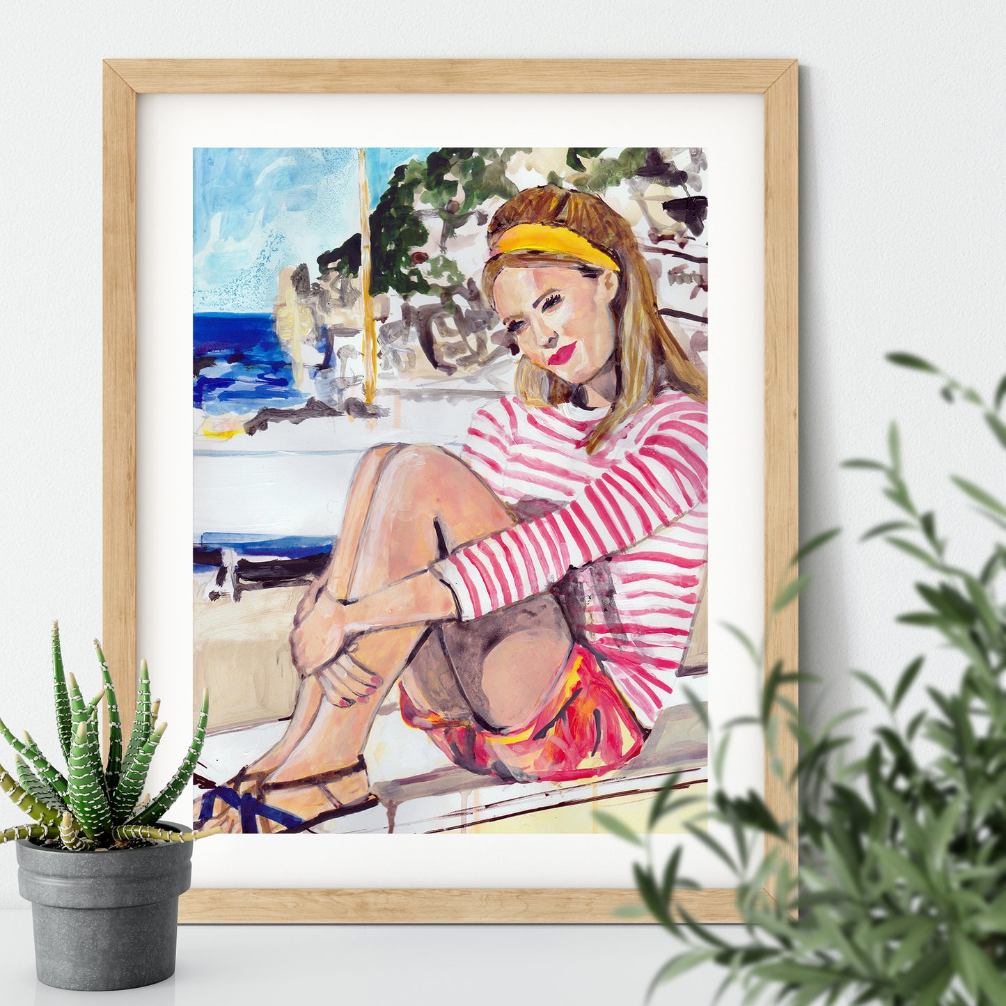 Image shows a framed illustration of a woman with a red and white striped top, red shorts and a yellow headband sitting on a wall by the coast with rocks and a blue sea in the background.