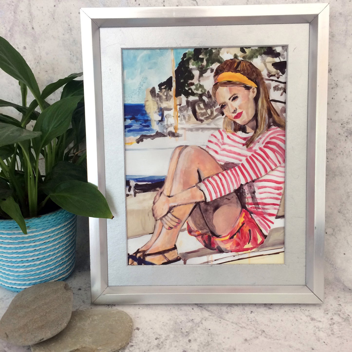 Image shows a framed illustration of a woman with a red and white striped top, red shorts and a yellow headband sitting on a wall by the coast with rocks and a blue sea in the background.