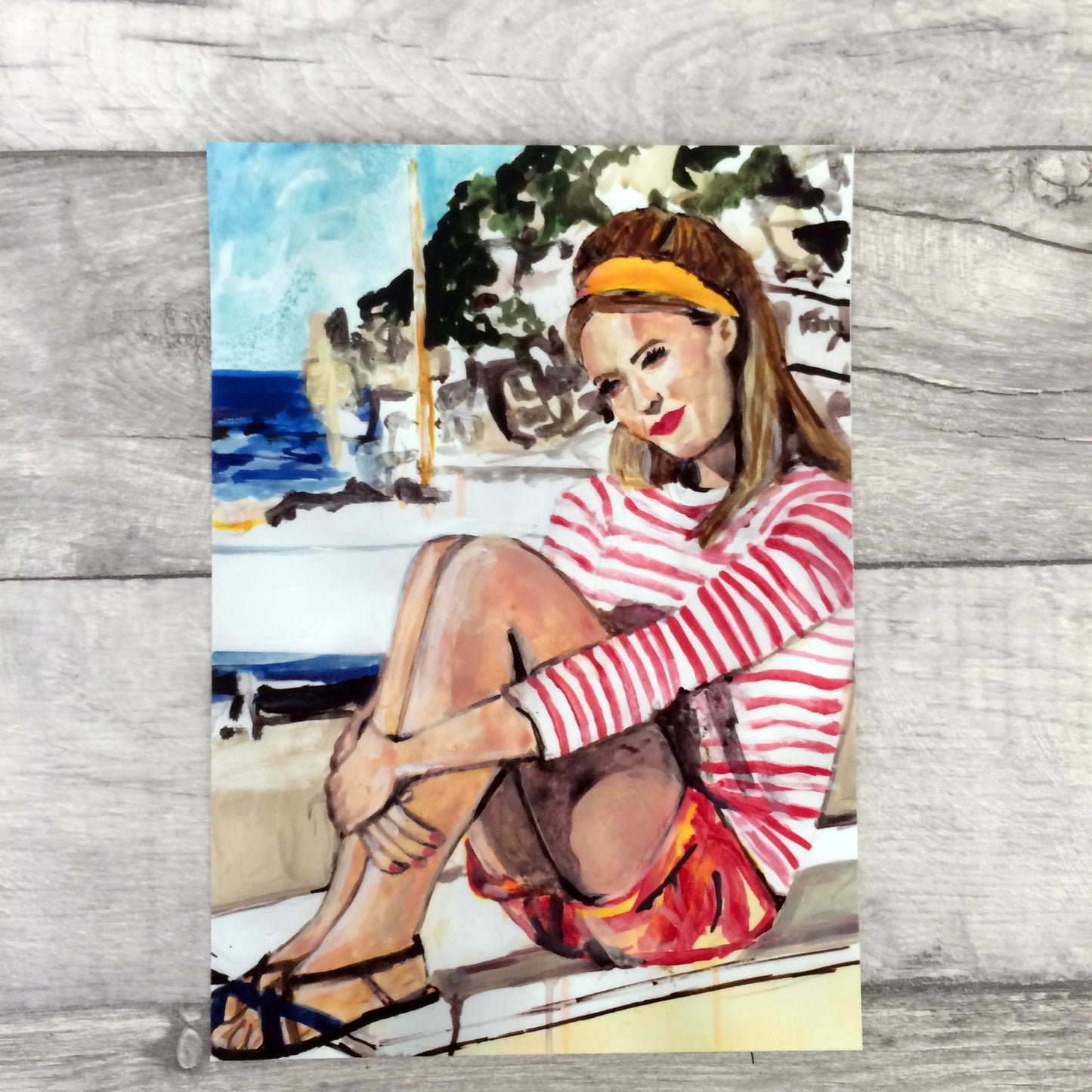 Image shows a framed illustration of a woman with a red and white striped top, red shorts and a yellow headband sitting on a wall by the coast with rocks and a blue sea in the background.