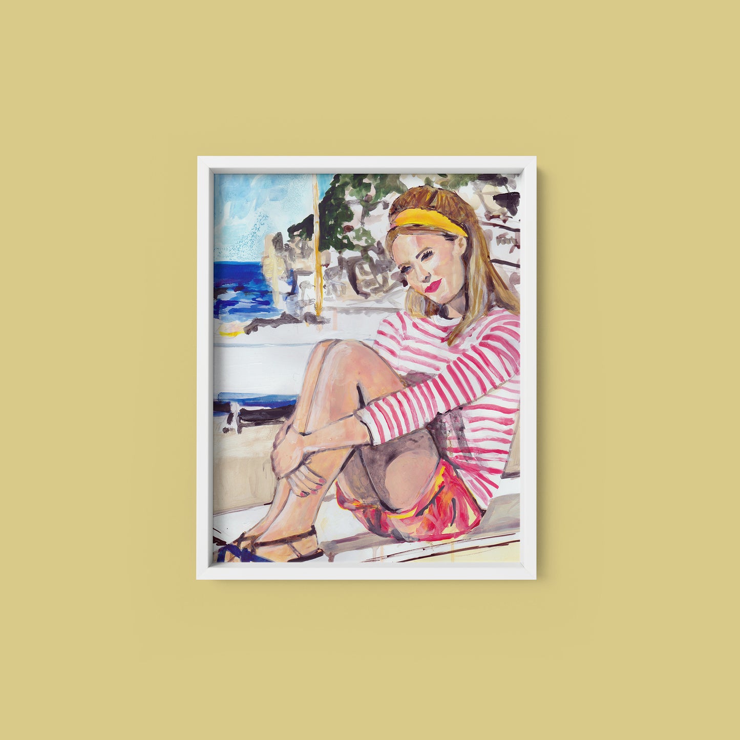 Image shows a framed illustration of a woman with a red and white striped top, red shorts and a yellow headband sitting on a wall by the coast with rocks and a blue sea in the background.