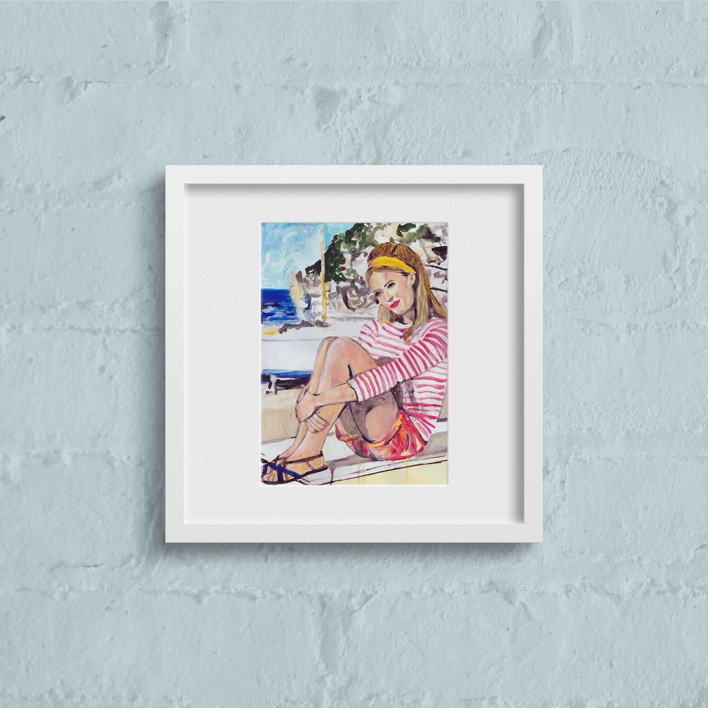Image shows a framed illustration of a woman with a red and white striped top, red shorts and a yellow headband sitting on a wall by the coast with rocks and a blue sea in the background.