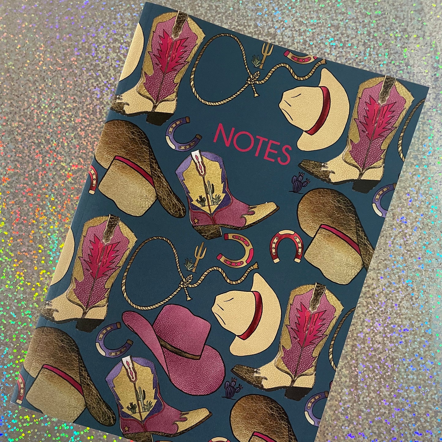Image of a blue notebook with a western inspired pattern of illustrated cowboy boots, hats, horseshoes and lassos with the word Notes in pink writing.