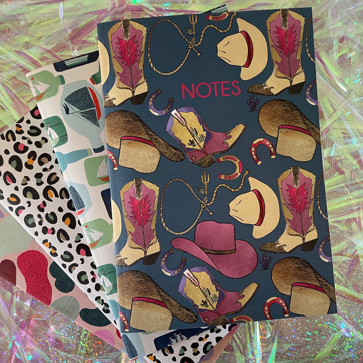 Image of a blue notebook with a western inspired pattern of illustrated cowboy boots, hats, horseshoes and lassos with the word Notes in pink writing.