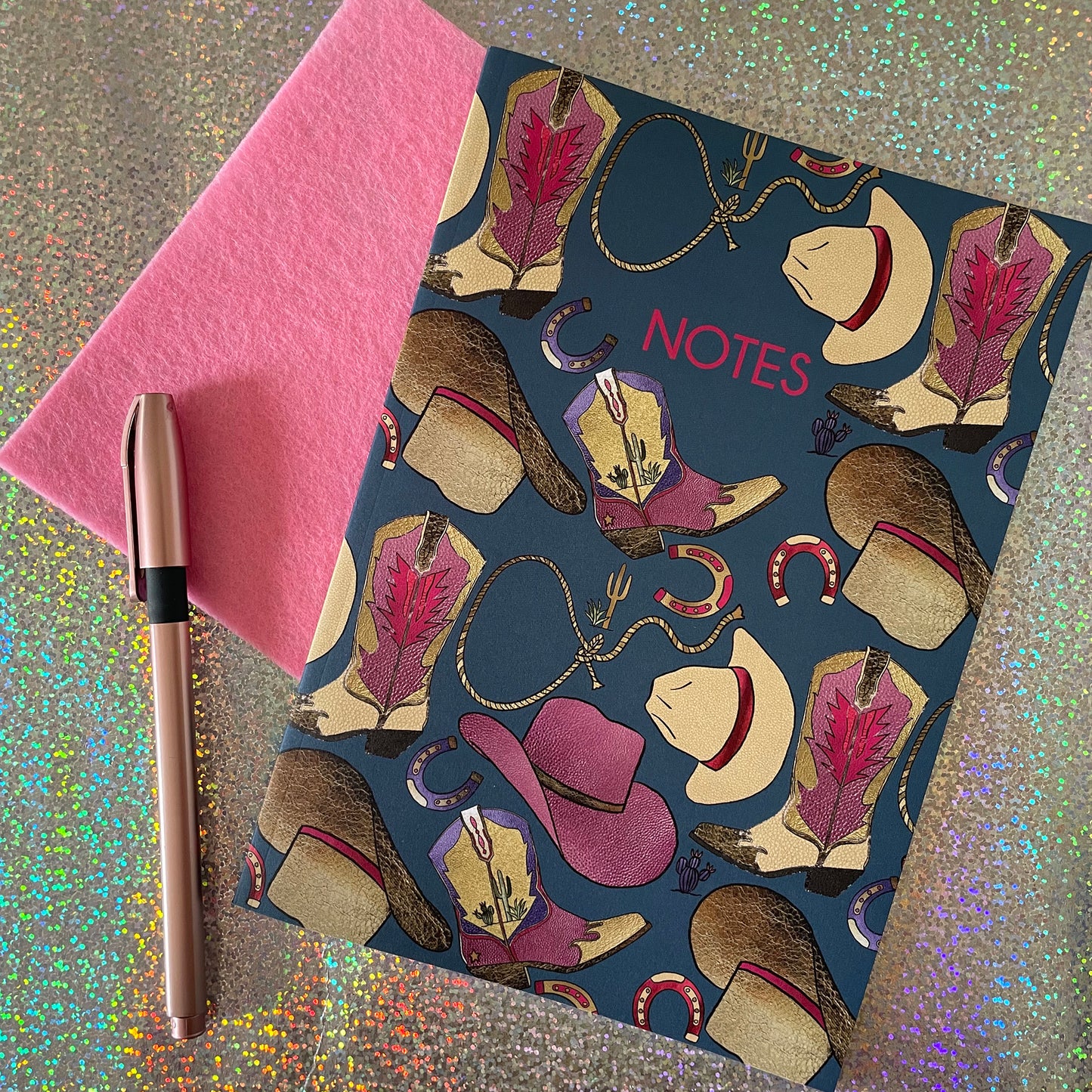 Image of a blue notebook with a western inspired pattern of illustrated cowboy boots, hats, horseshoes and lassos with the word Notes in pink writing.