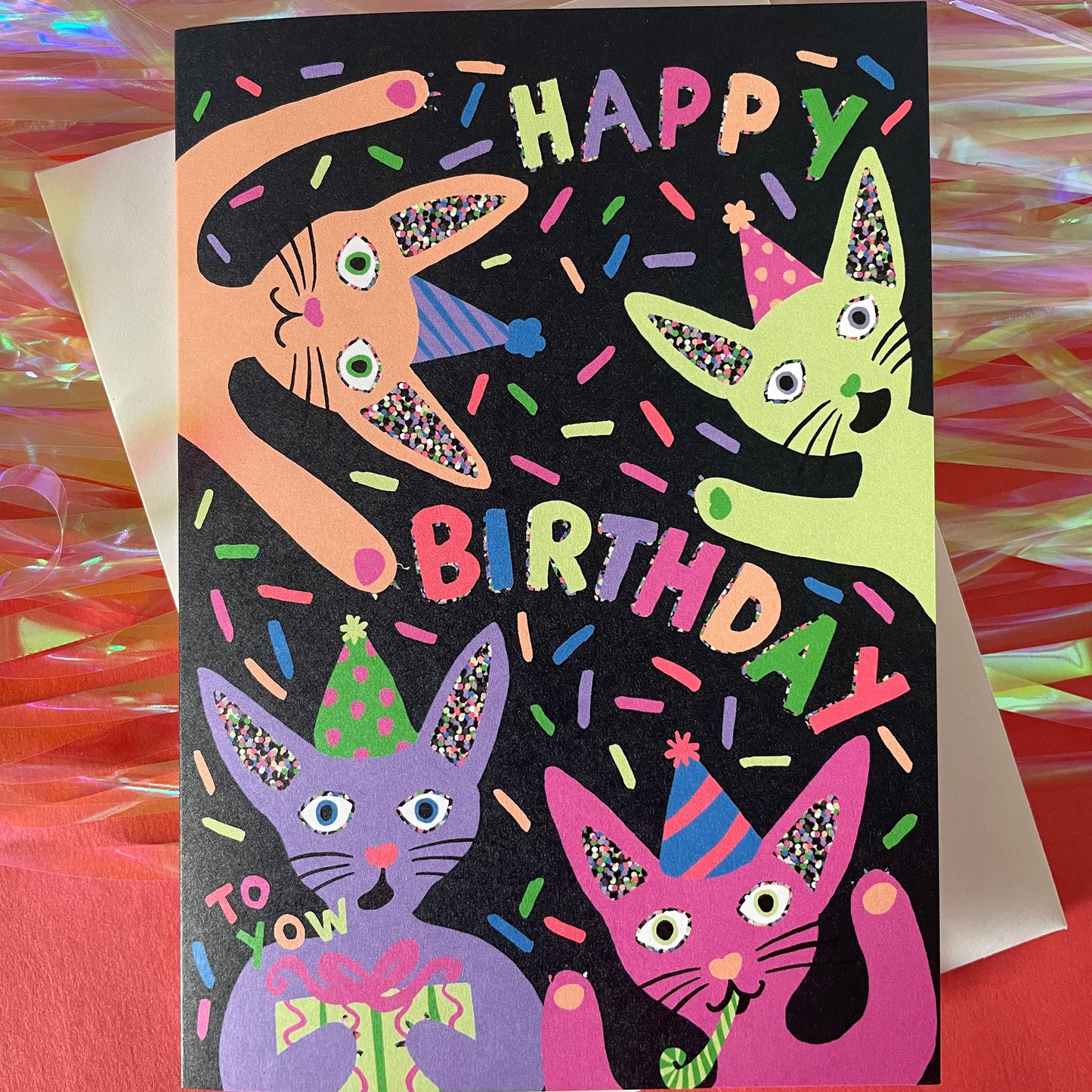 Image shows a quirky black greetings card with neon, illustrated cats in party hats and the words happy birthday.