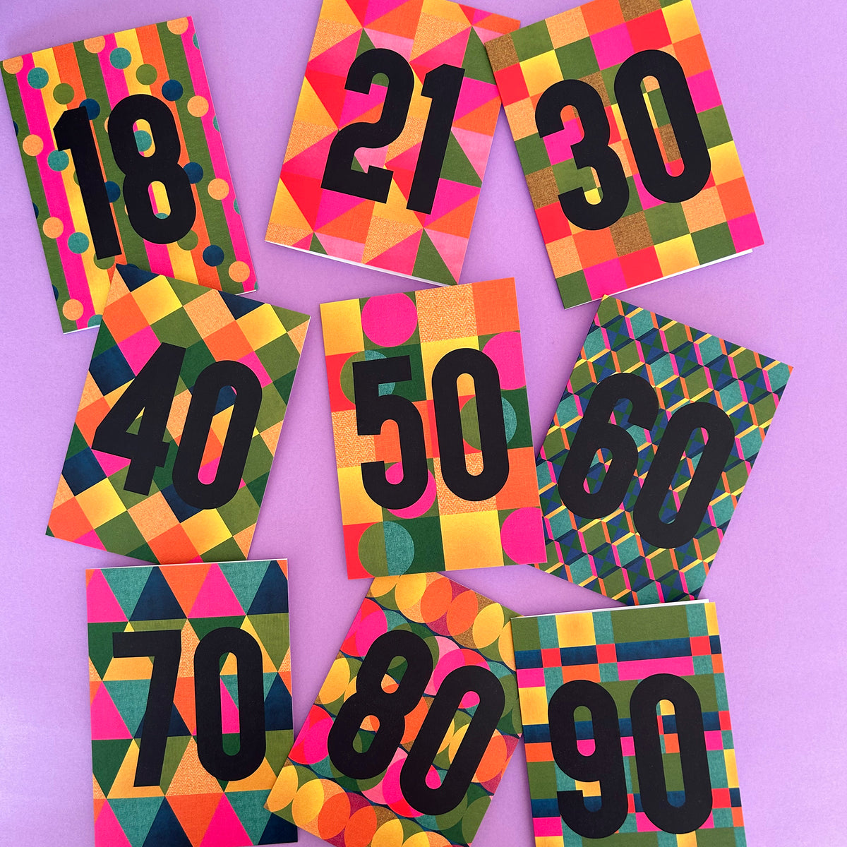 A set of colourful numbered cards or tags arranged on a lavender background, with numbers 18 through 90 printed in large black font over geometric patterned designs in pink, yellow, green, orange, and blue.