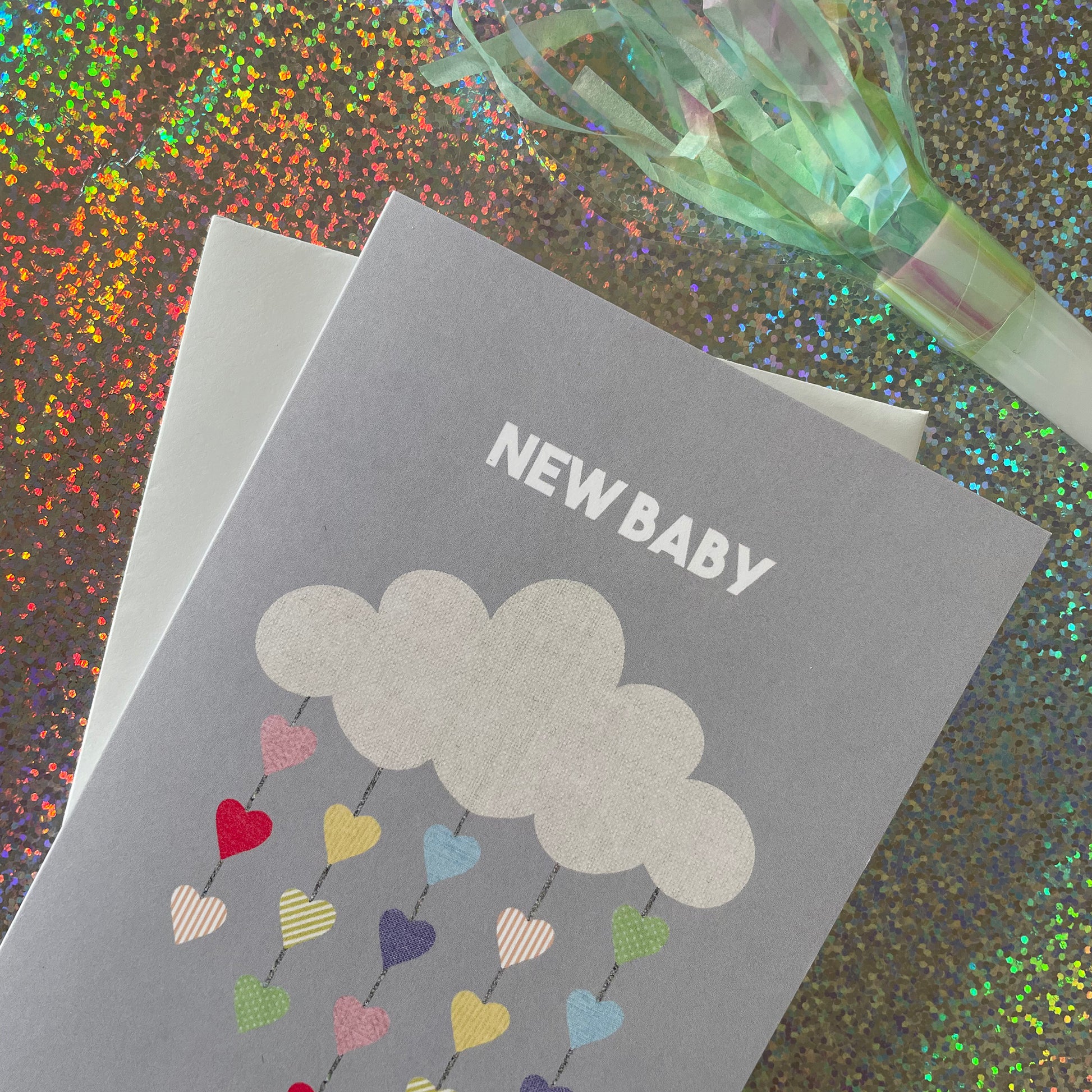 Image shows a pale grey greetings card with an illustration of a cloud with colourful hearts hanging from it and the words 'New Baby'
