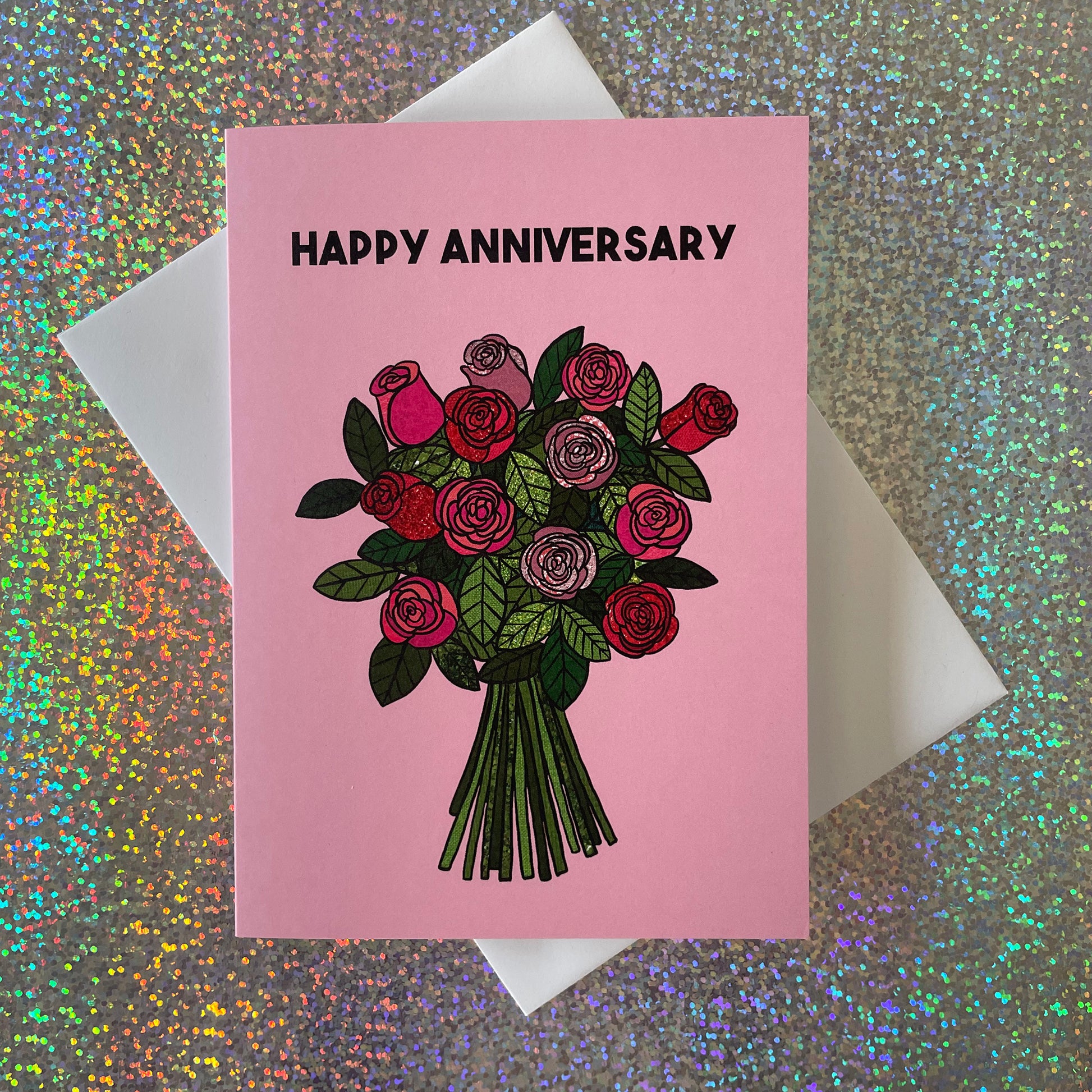 Image shows a light pink greetings card with an illustration of a bunch of roses and the words 'Happy Anniversary'