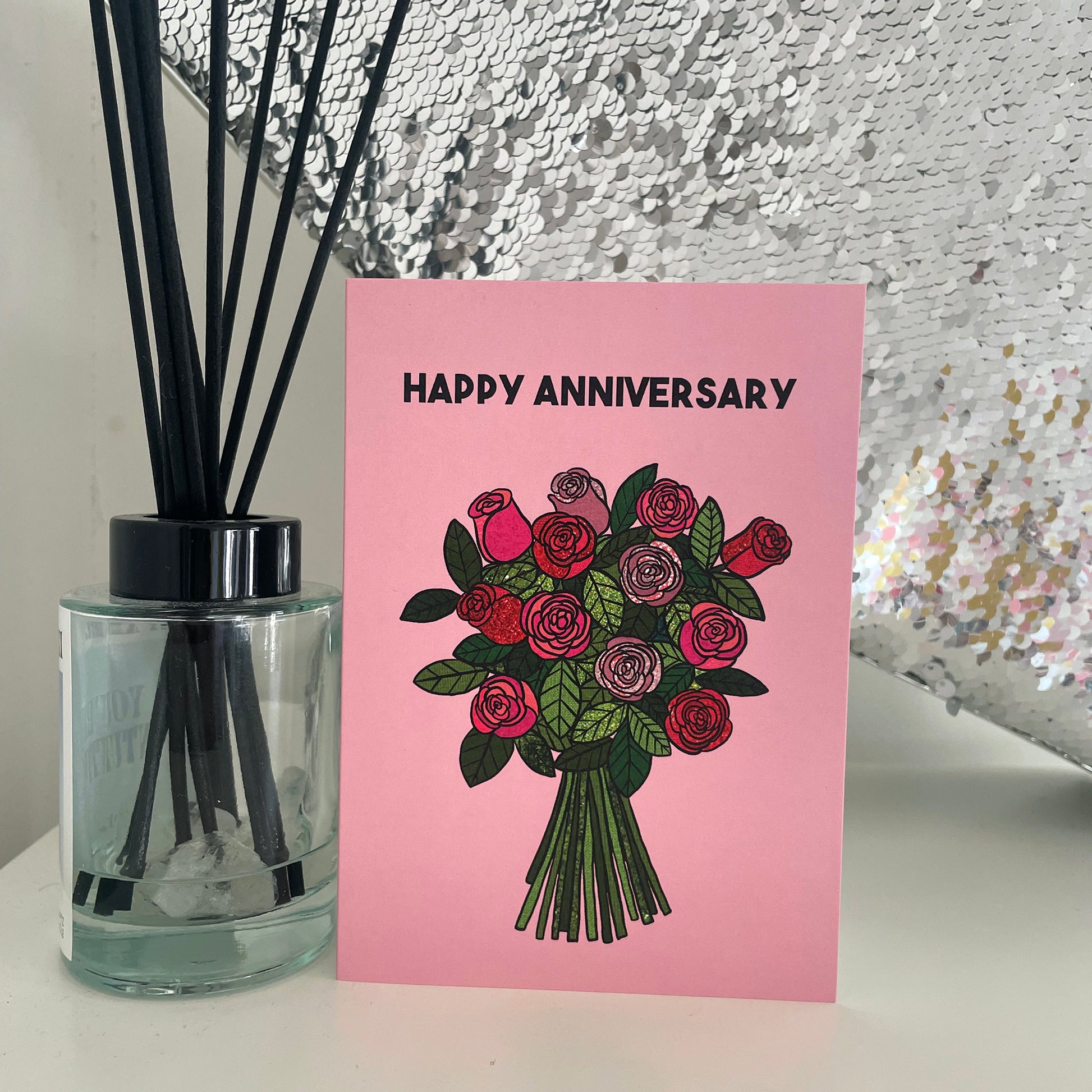 Image shows a light pink greetings card with an illustration of a bunch of roses and the words 'Happy Anniversary'