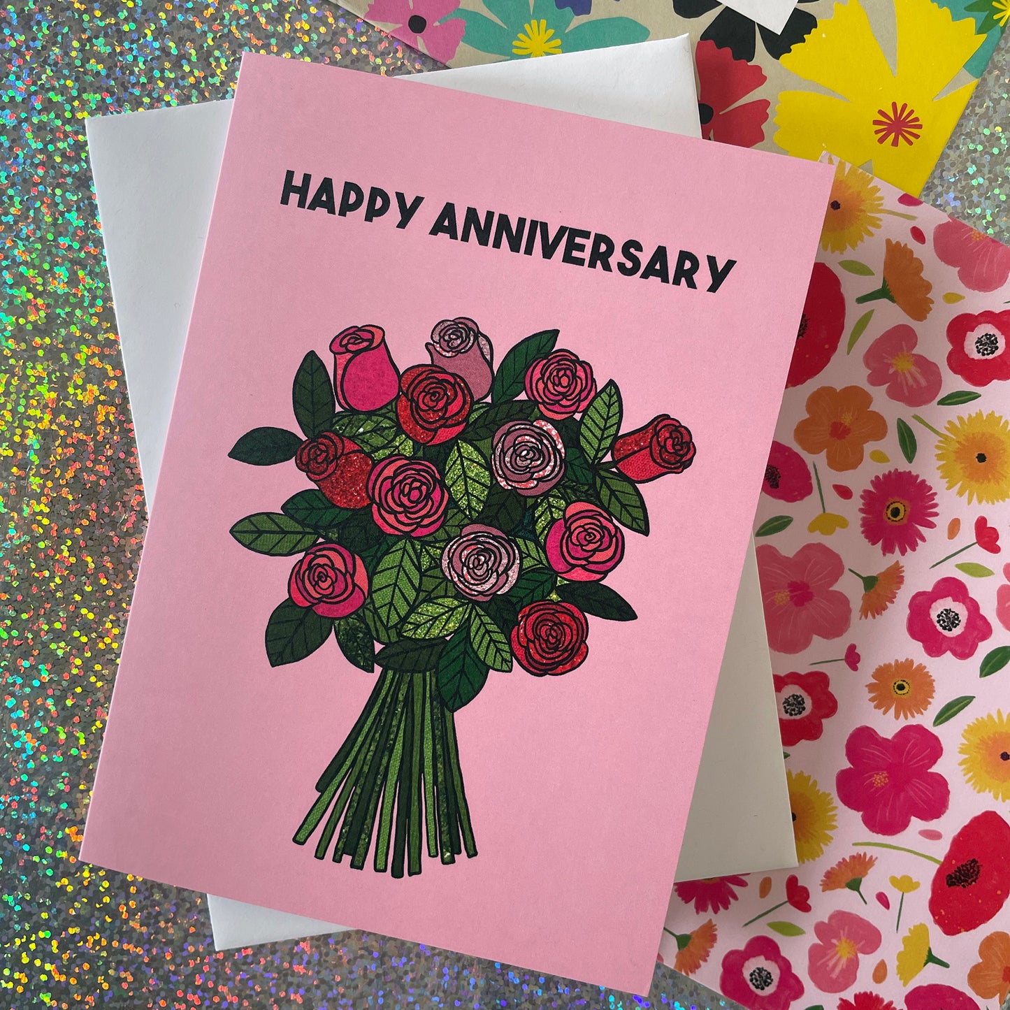 Image shows a light pink greetings card with an illustration of a bunch of roses and the words 'Happy Anniversary'