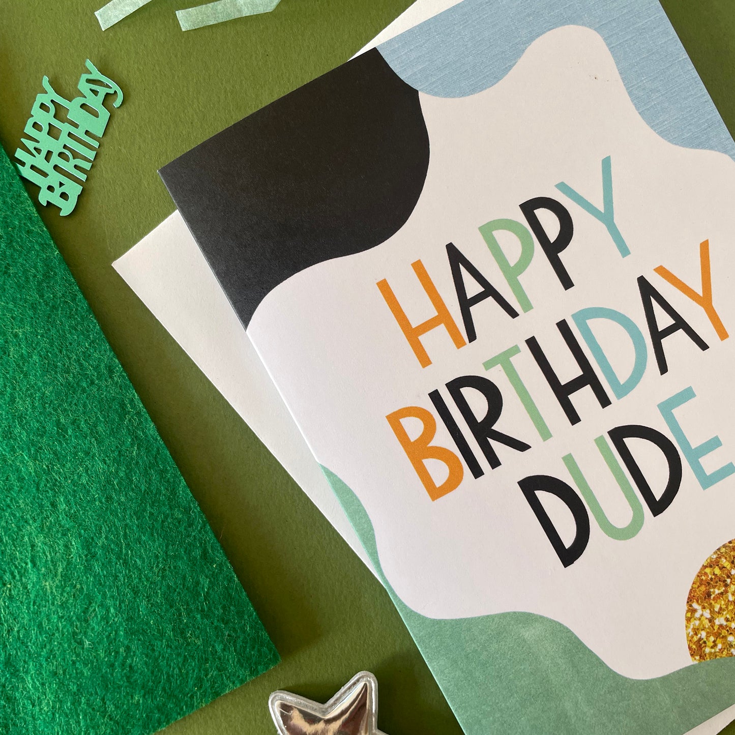 Image shows a white greetings crad with pale blue, green, black and gold abstract shapes and the words 'Happy Birthday Dude'