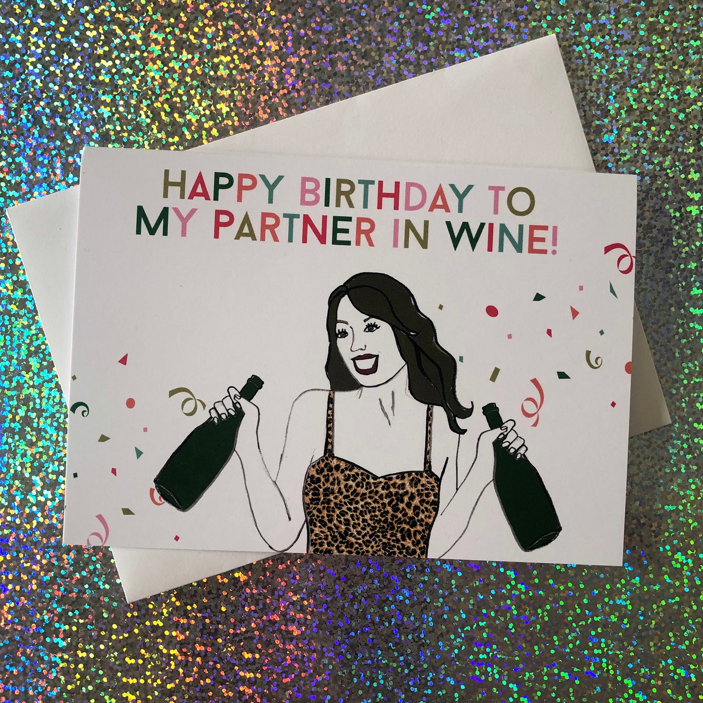 Image shows a fun and fabulous white birthday card with an illustration of a woman in a leopard print top holding 2 bottles of wine and the wording 'Happy Birthday to my partner in wine'..