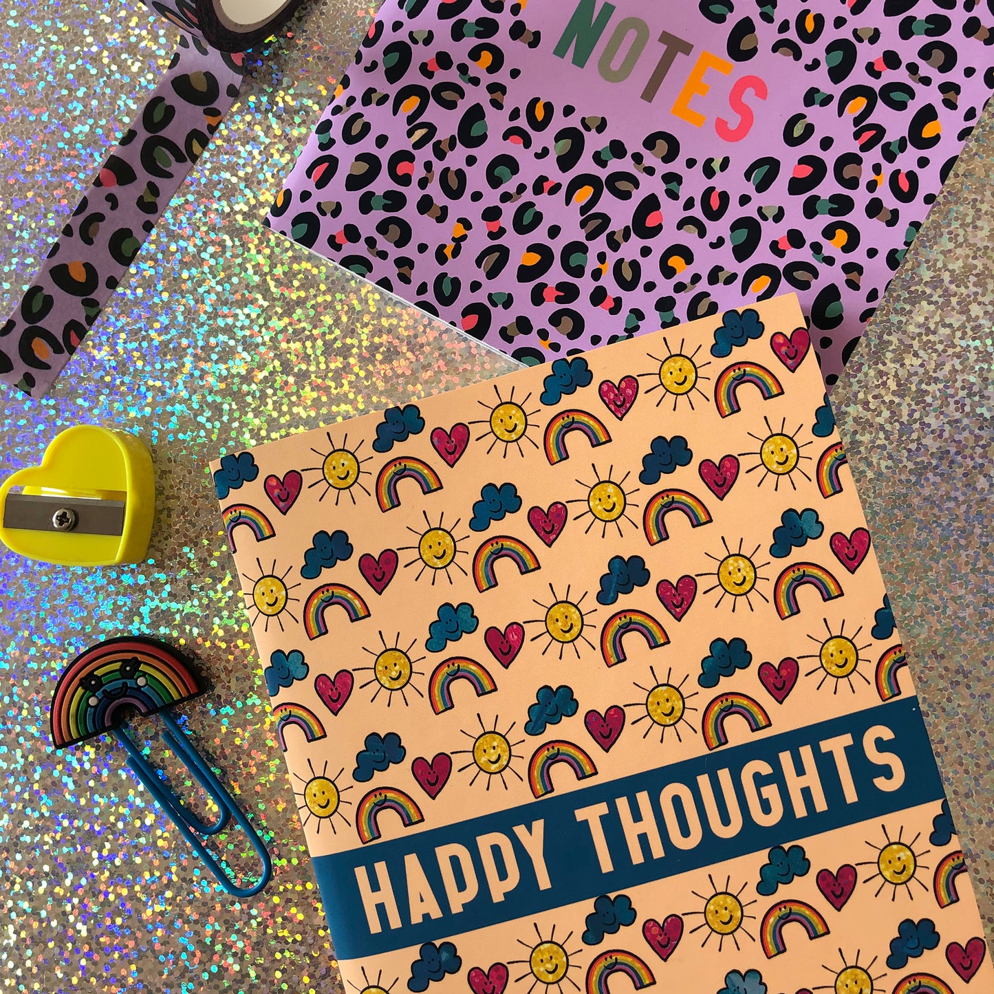 Image shows a patterned notebook with illustrations of suns, clouds, hearts and rainbows with smiling faces. The notebook says Happy thoughts on the cover