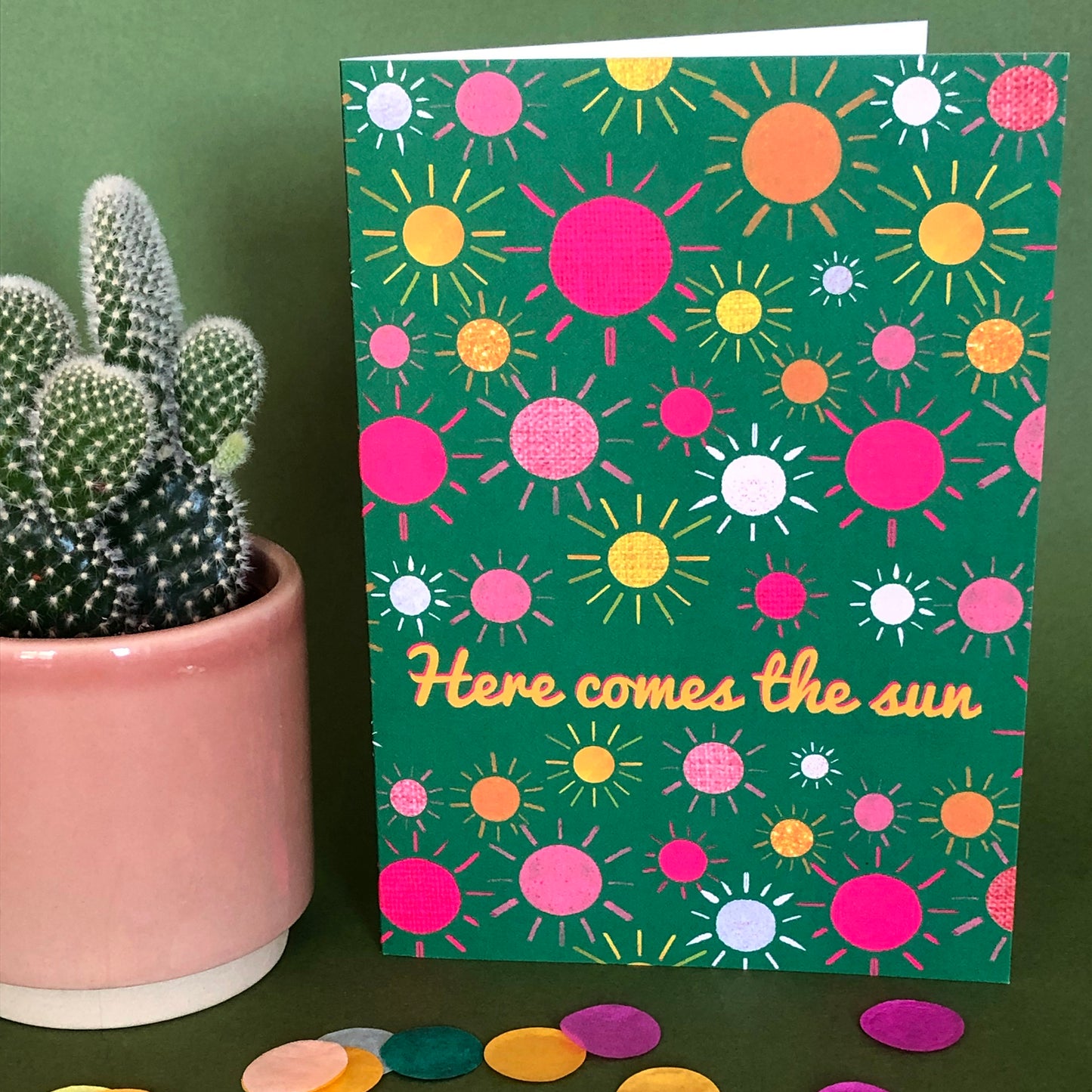 Image shows a green everyday greetings card that says 'here comes the sun' and features a fun pattern of colourful suns