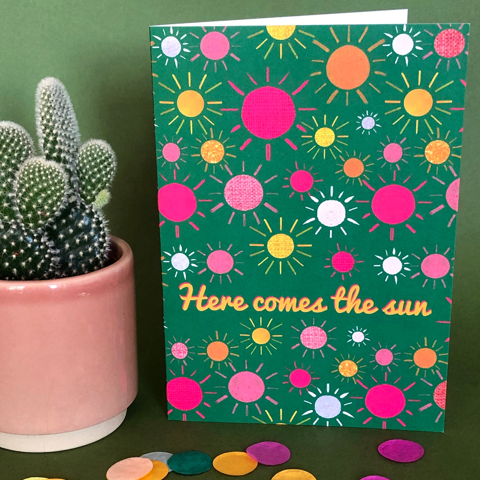 Image shows a green everyday greetings card that says 'here comes the sun' and features a fun pattern of colourful suns
