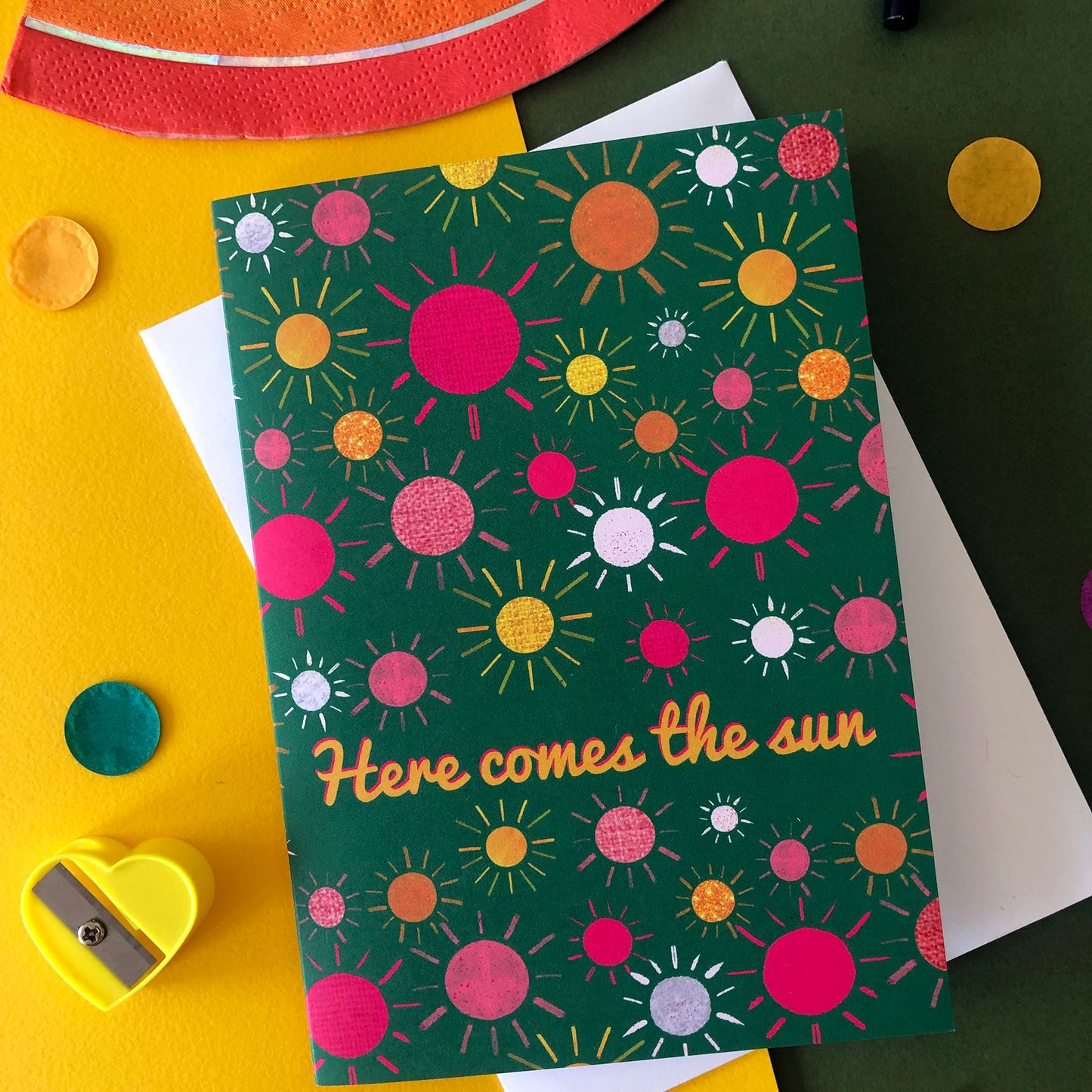 Image shows a green everyday greetings card that says 'here comes the sun' and features a fun pattern of colourful suns