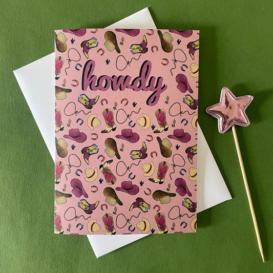 Image of a dusty pink coloured greetings card that features an illustrated pattern of cowboy boots, hats, horseshoes and lassos. It says "Howdy" in the middle.