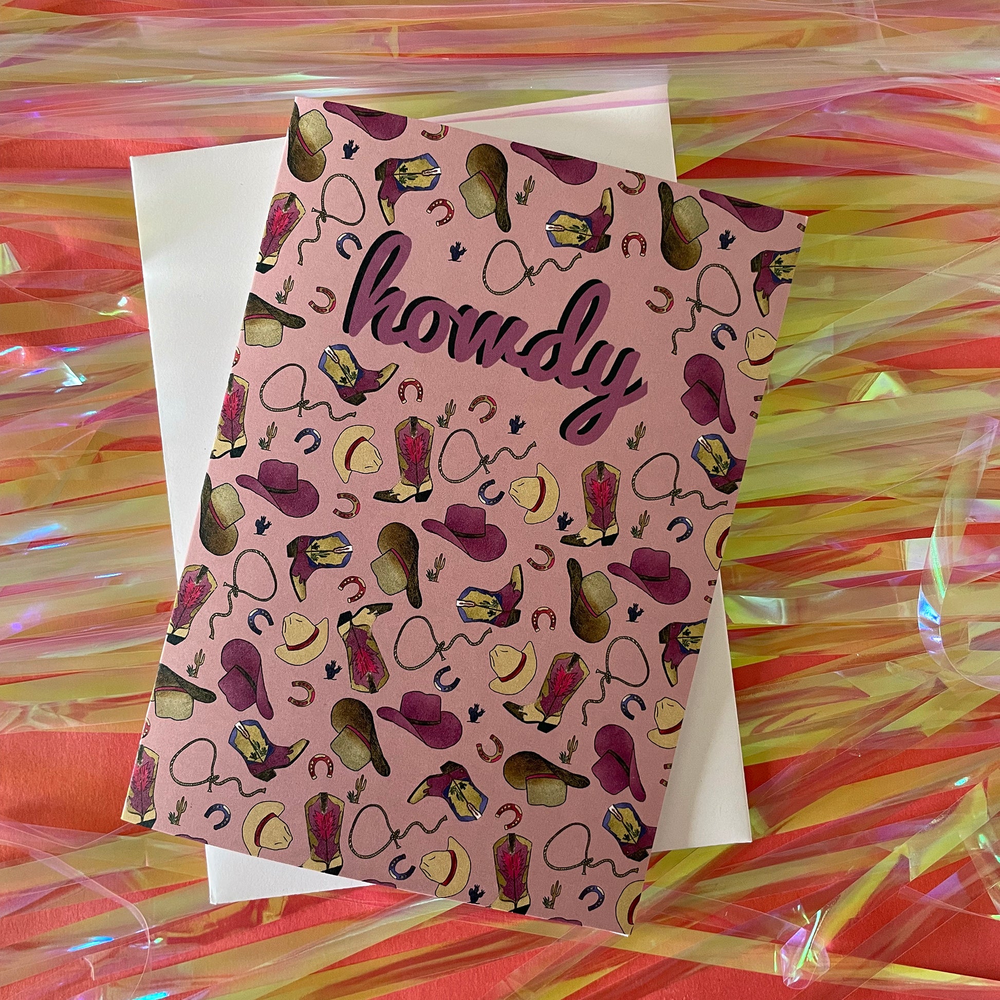 Image of a dusty pink coloured greetings card that features an illustrated pattern of cowboy boots, hats, horseshoes and lassos. It says "Howdy" in the middle.