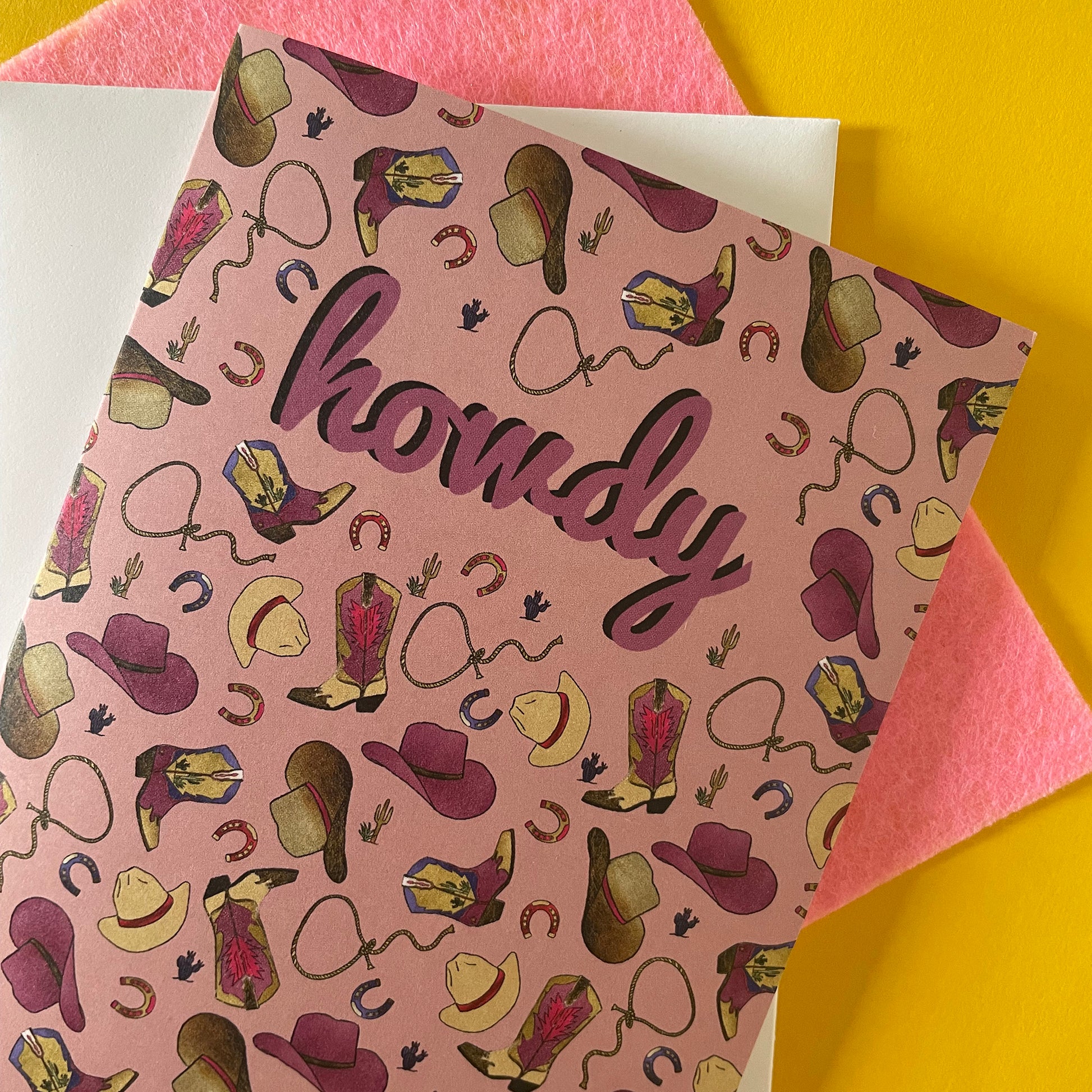 Close up image of a dusty pink coloured greetings card that features an illustrated pattern of cowboy boots, hats, horseshoes and lassos. It says "Howdy" in the middle.