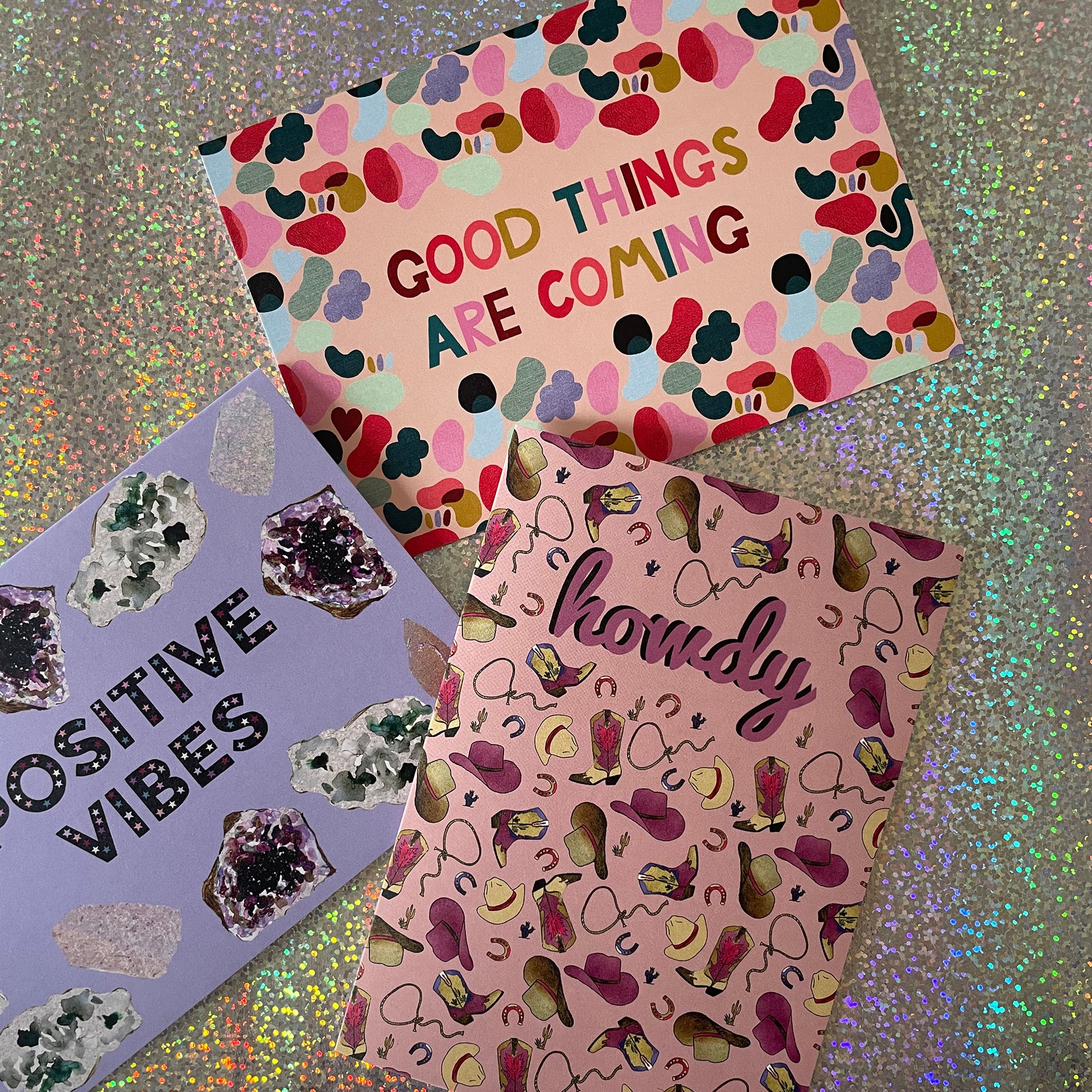 Image of 3 cards, a bright and colourful card with an abstract shape design and the words 'Good Things Are Coming', a lavender coloured card with illustrations of crystals and the words 'Positive Vibes' and a pale pink card with a western inspired pattern featuring hats, boots and lassos that says 'Howdy"