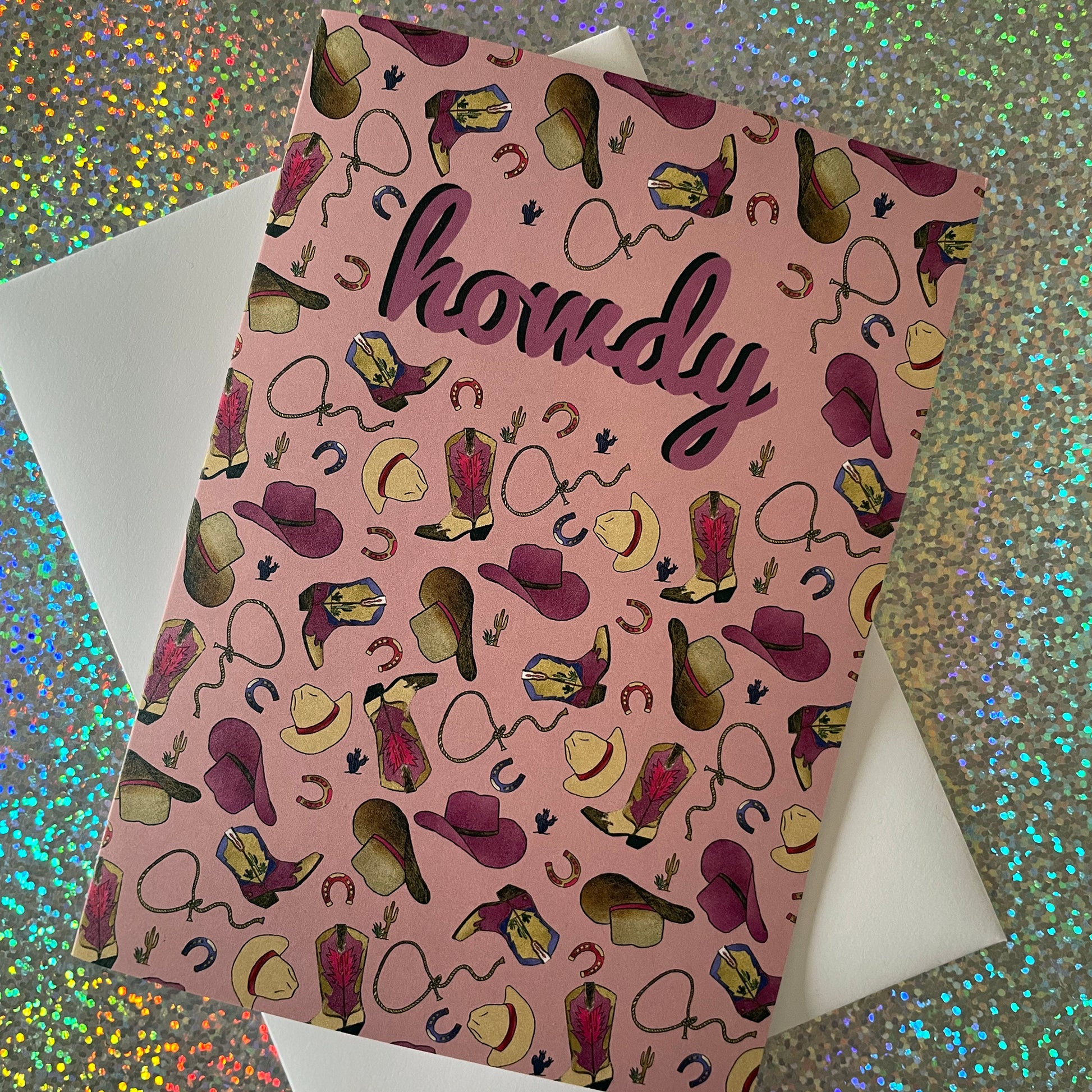 Image of a dusty pink coloured greetings card that features an illustrated pattern of cowboy boots, hats, horseshoes and lassos. It says "Howdy" in the middle.