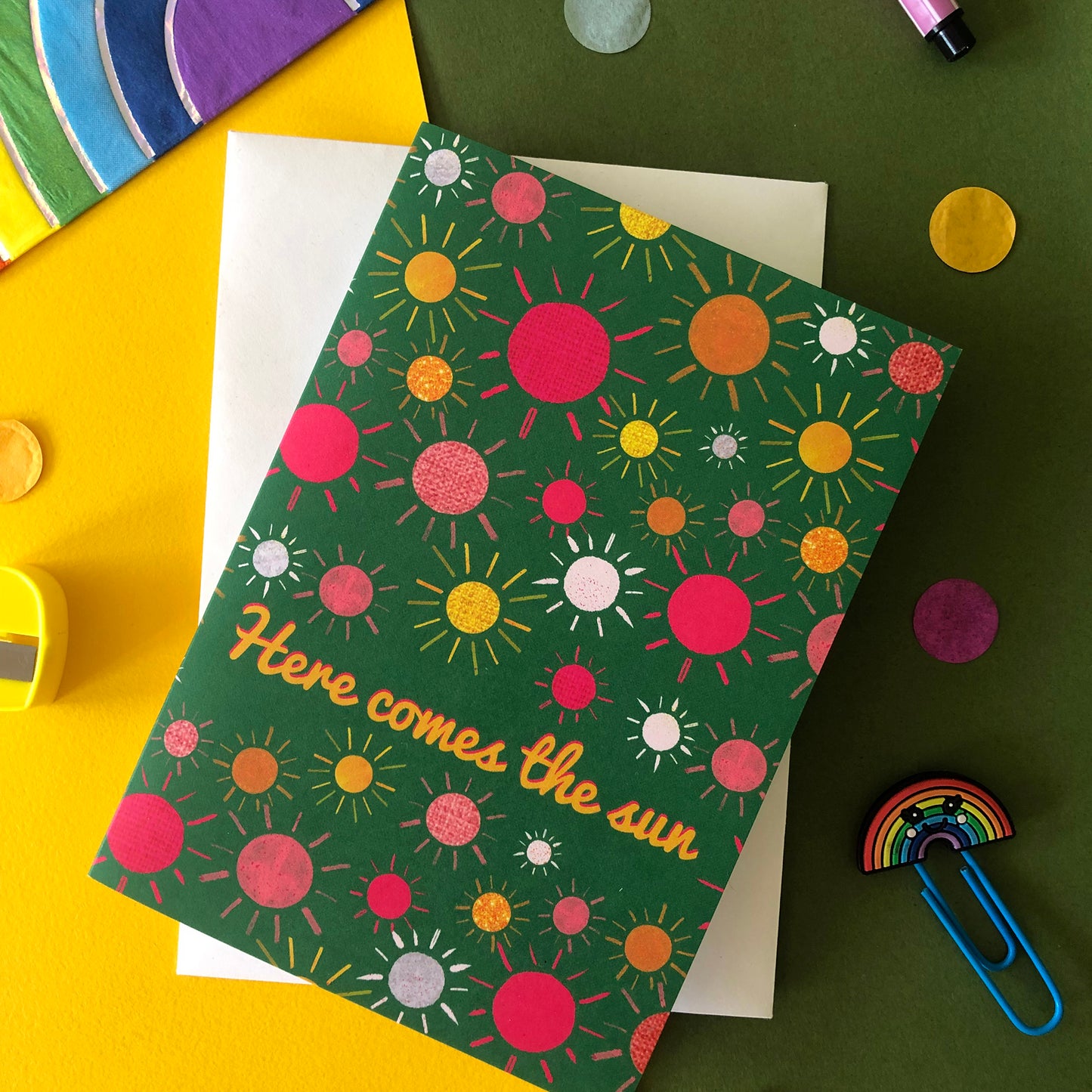 Image shows a green everyday greetings card that says 'here comes the sun' and features a fun pattern of colourful suns