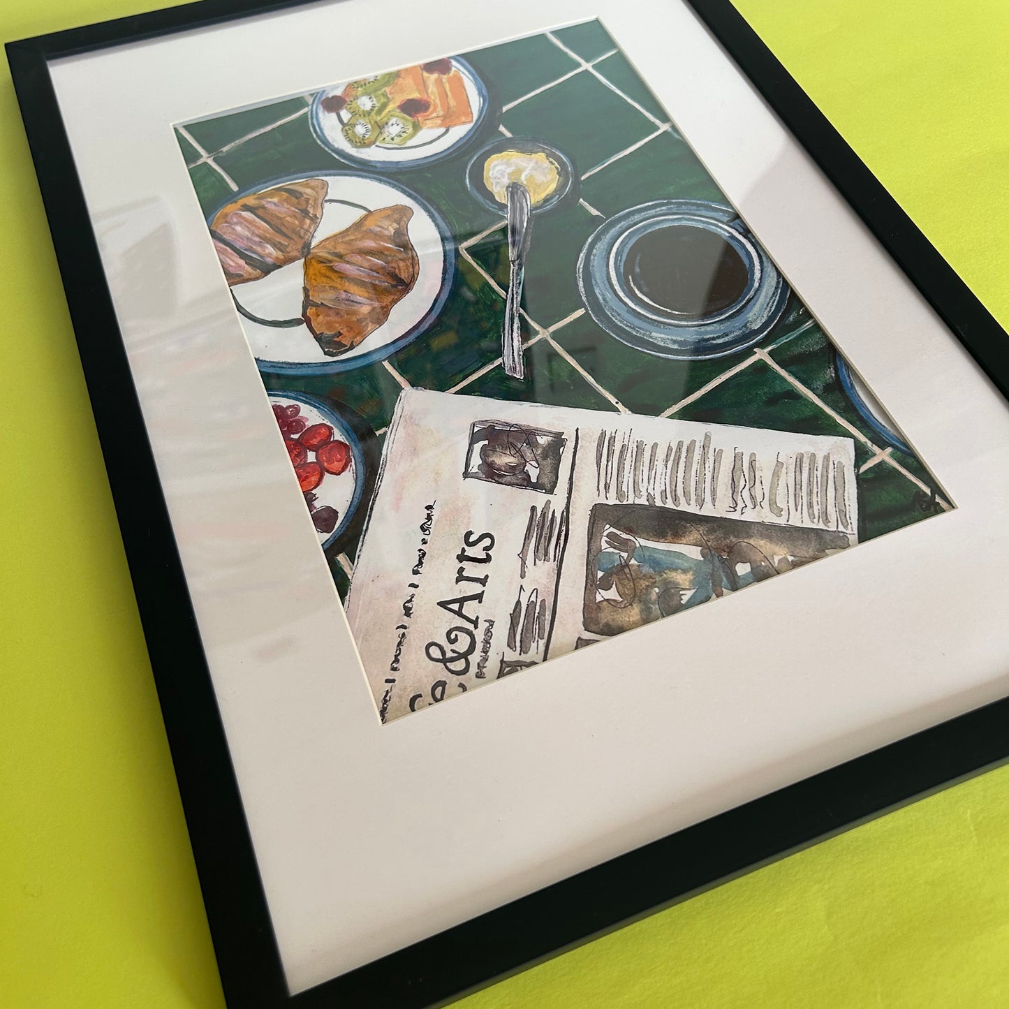 A framed art print featuring a still life illustration of a breakfast table with croissants, plates, a newspaper, and a glass of milk on a green tiled background, displayed on a bright yellow surface.