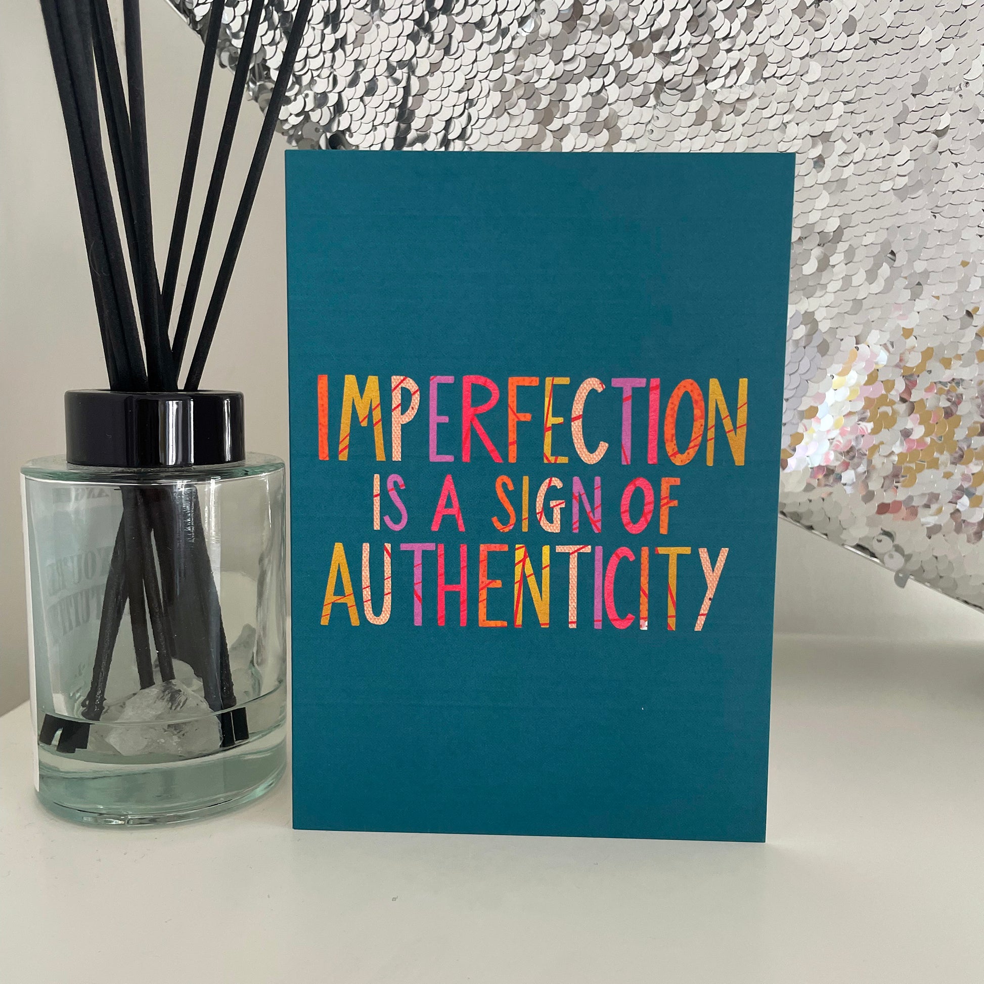 Image shows a teal coloured greetings card with the wording 'Imperfection is a sign of authenticity'