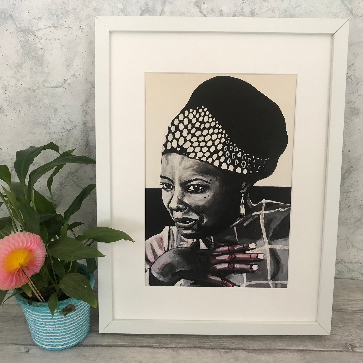 Image shows a framed black and white portrait of Maya Angelou. Artwork is painted in black and white wirth a pink undertone visible in the hand and shoulder