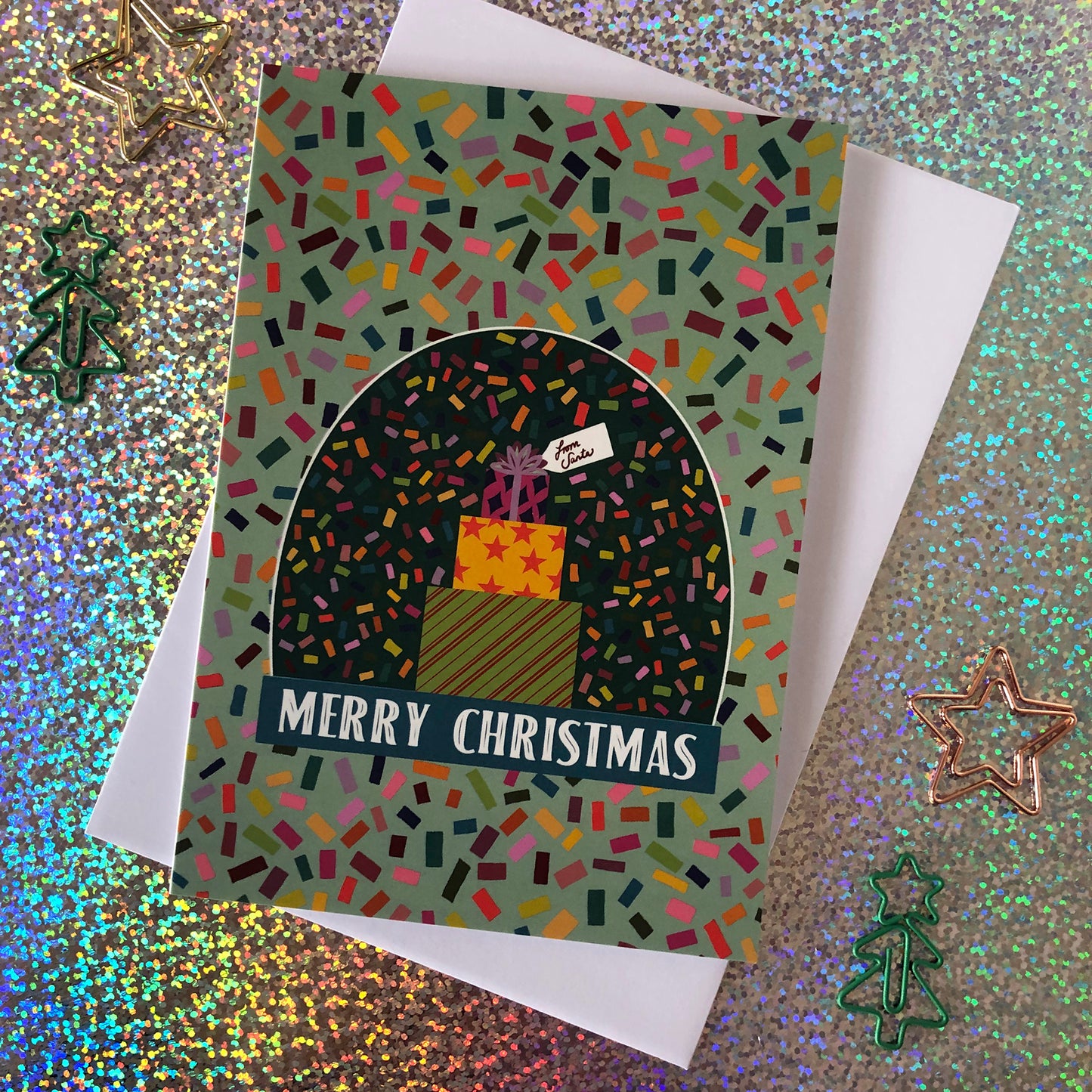 Image shows a bright and colourful Christmas card featuring an illustration of a snow globe on a confetti patterned background.