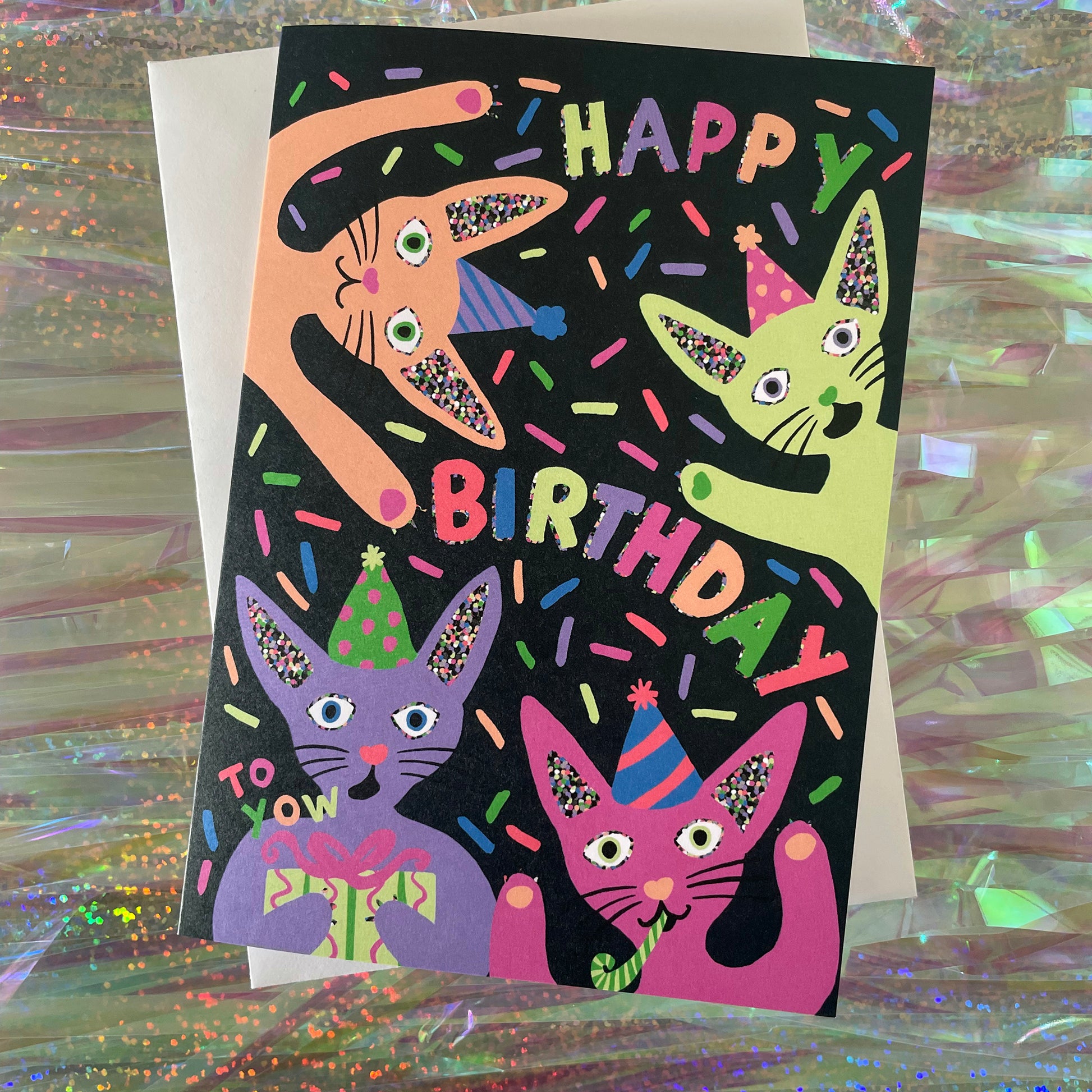 Image shows a quirky black greetings card with neon, illustrated cats in party hats and the words happy birthday.
