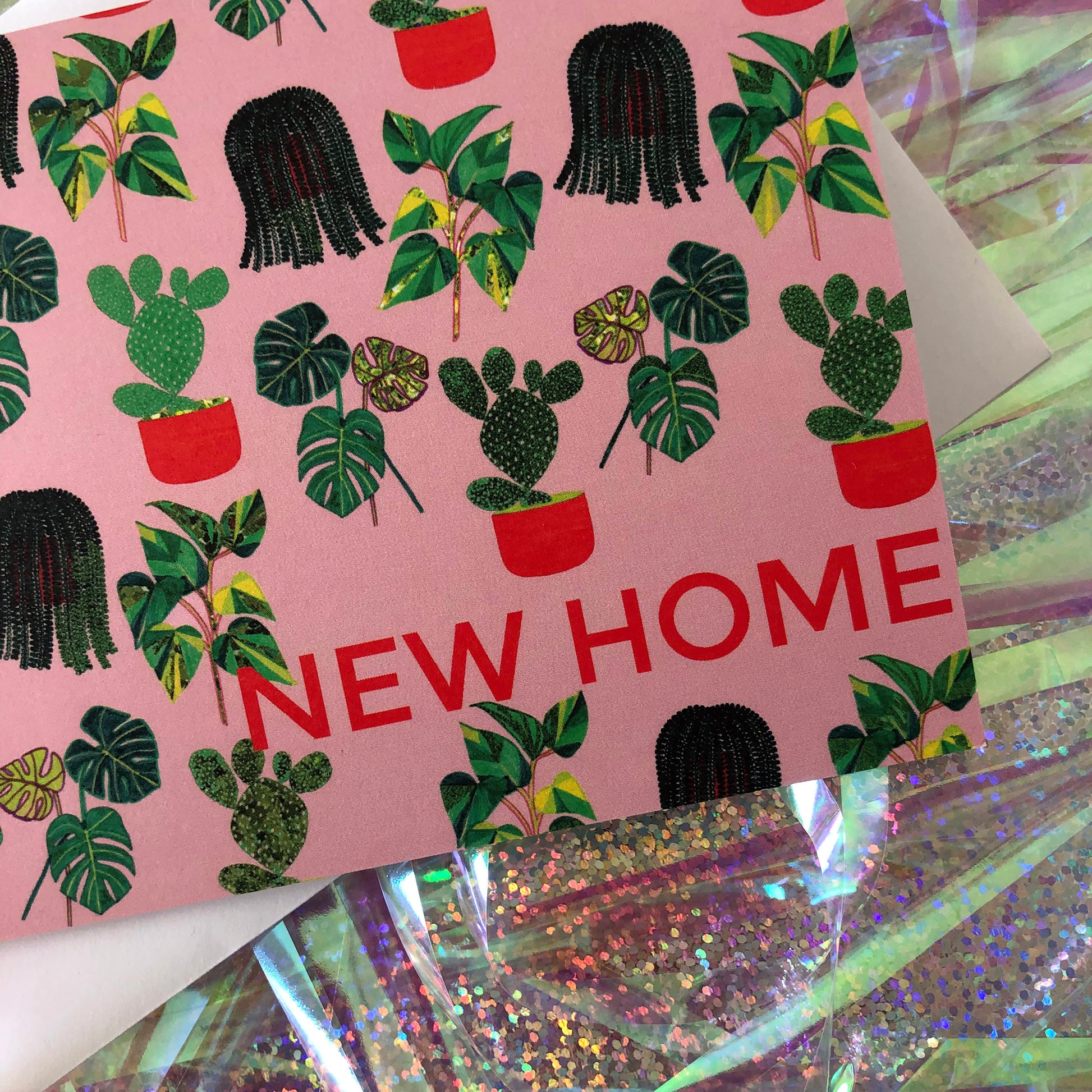 Image shows a pastel pink new home card with a pattern of illustrated green house plants and orange pots and writing.