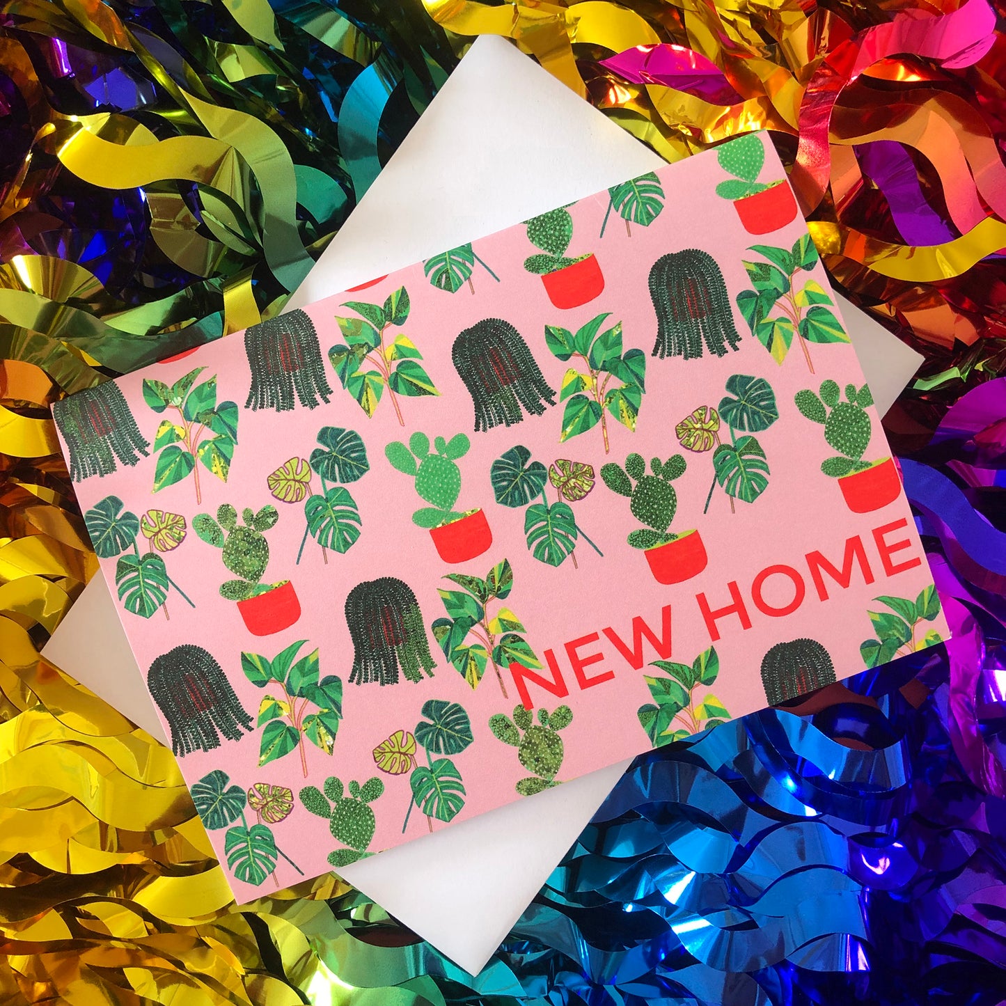 Image shows a pastel pink new home card with a pattern of illustrated green house plants and orange pots and writing.