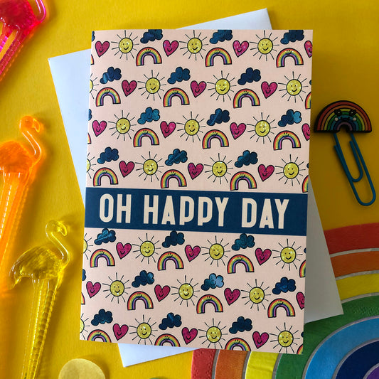 Image shows an everyday greetings card that says 'oh happy day' and features a fun pattern of suns, clouds and rainbows