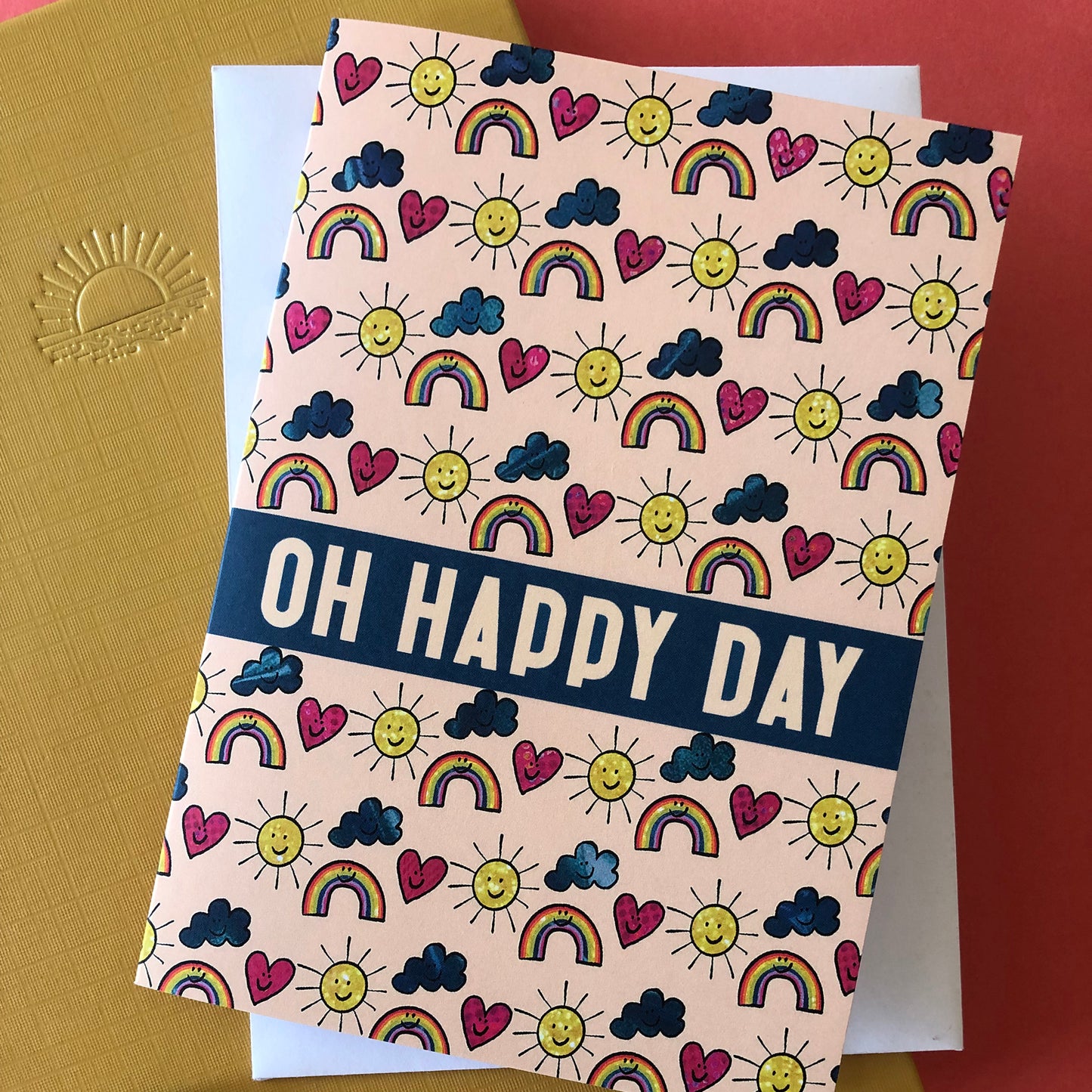 Image shows an everyday greetings card that says 'oh happy day' and features a fun pattern of suns, clouds and rainbows
