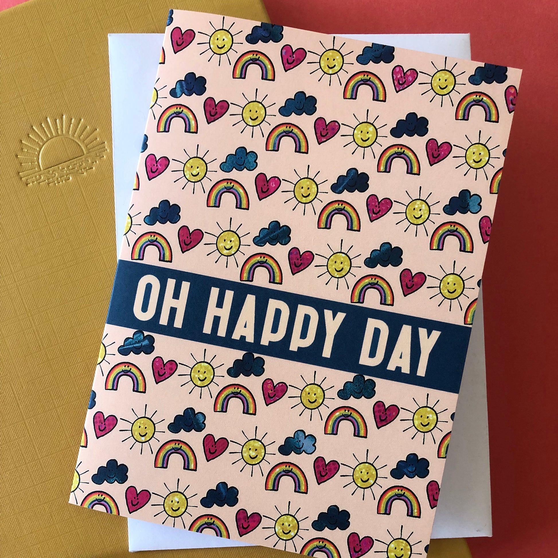 Image shows an everyday greetings card that says 'oh happy day' and features a fun pattern of suns, clouds and rainbows