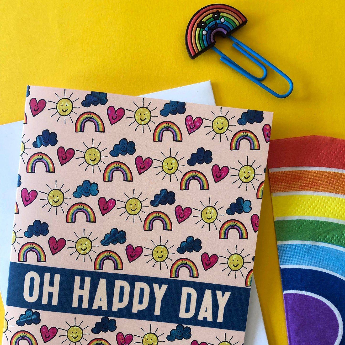 Image shows an everyday greetings card that says 'oh happy day' and features a fun pattern of suns, clouds and rainbows