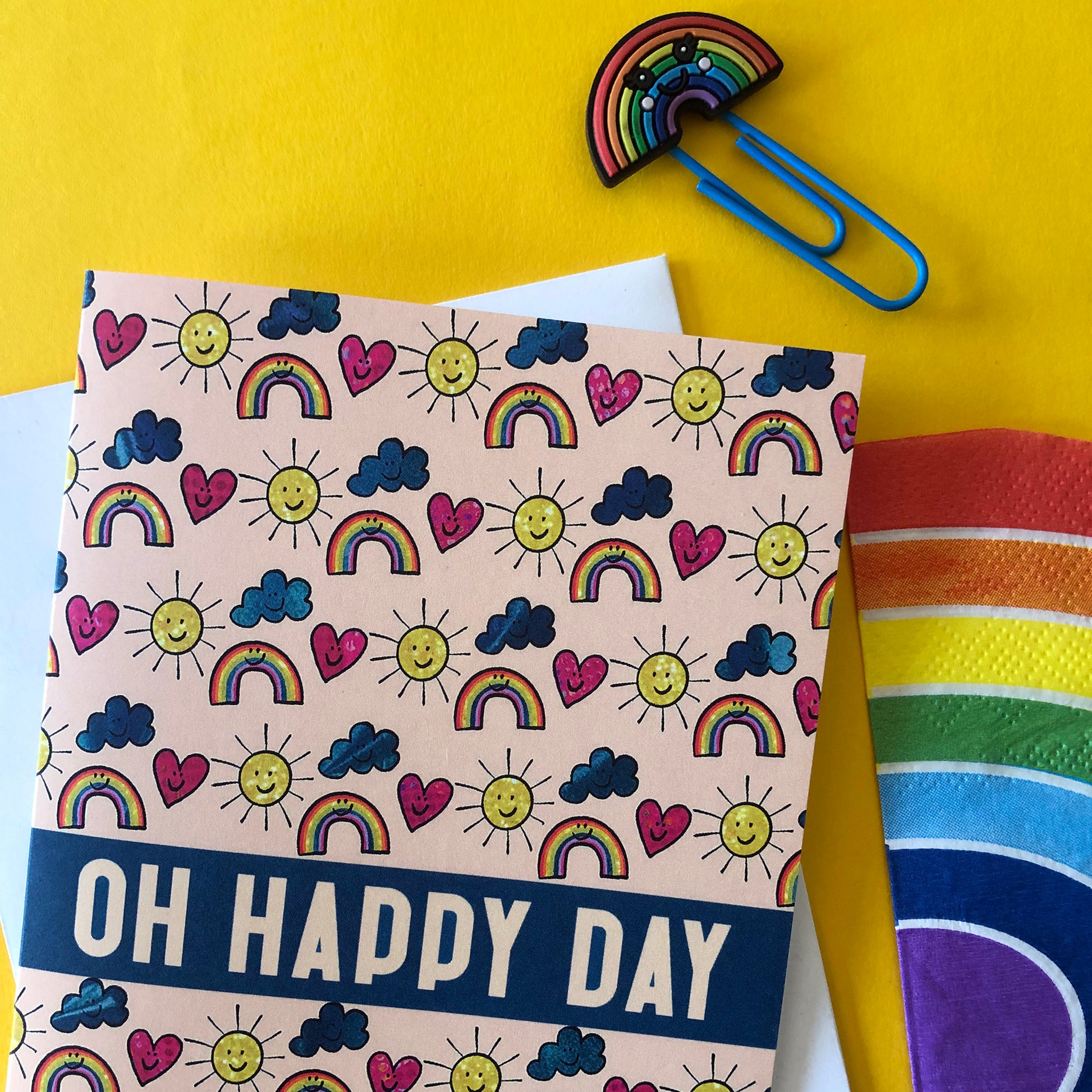 Image shows an everyday greetings card that says 'oh happy day' and features a fun pattern of suns, clouds and rainbows