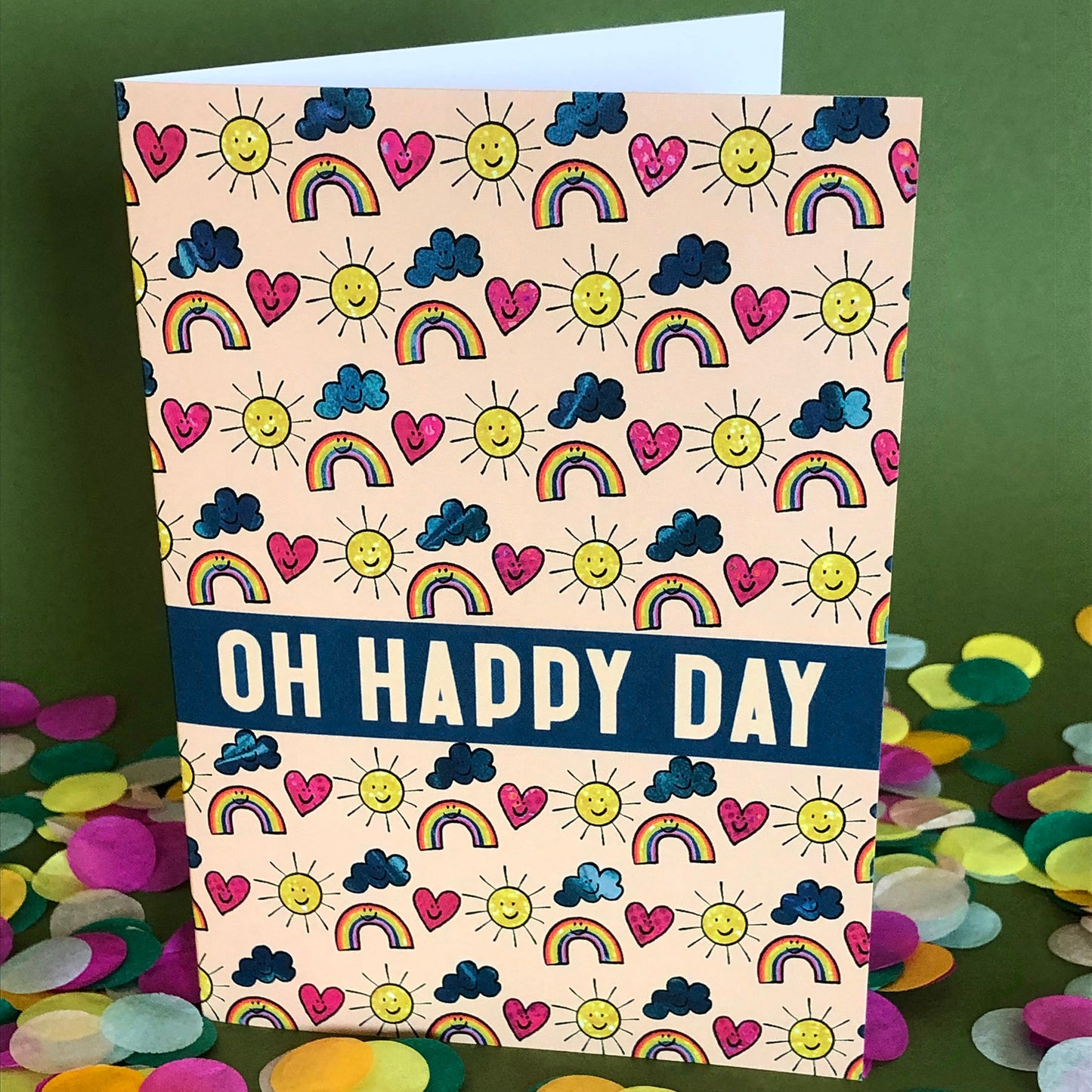 Image shows an everyday greetings card that says 'oh happy day' and features a fun pattern of suns, clouds and rainbows
