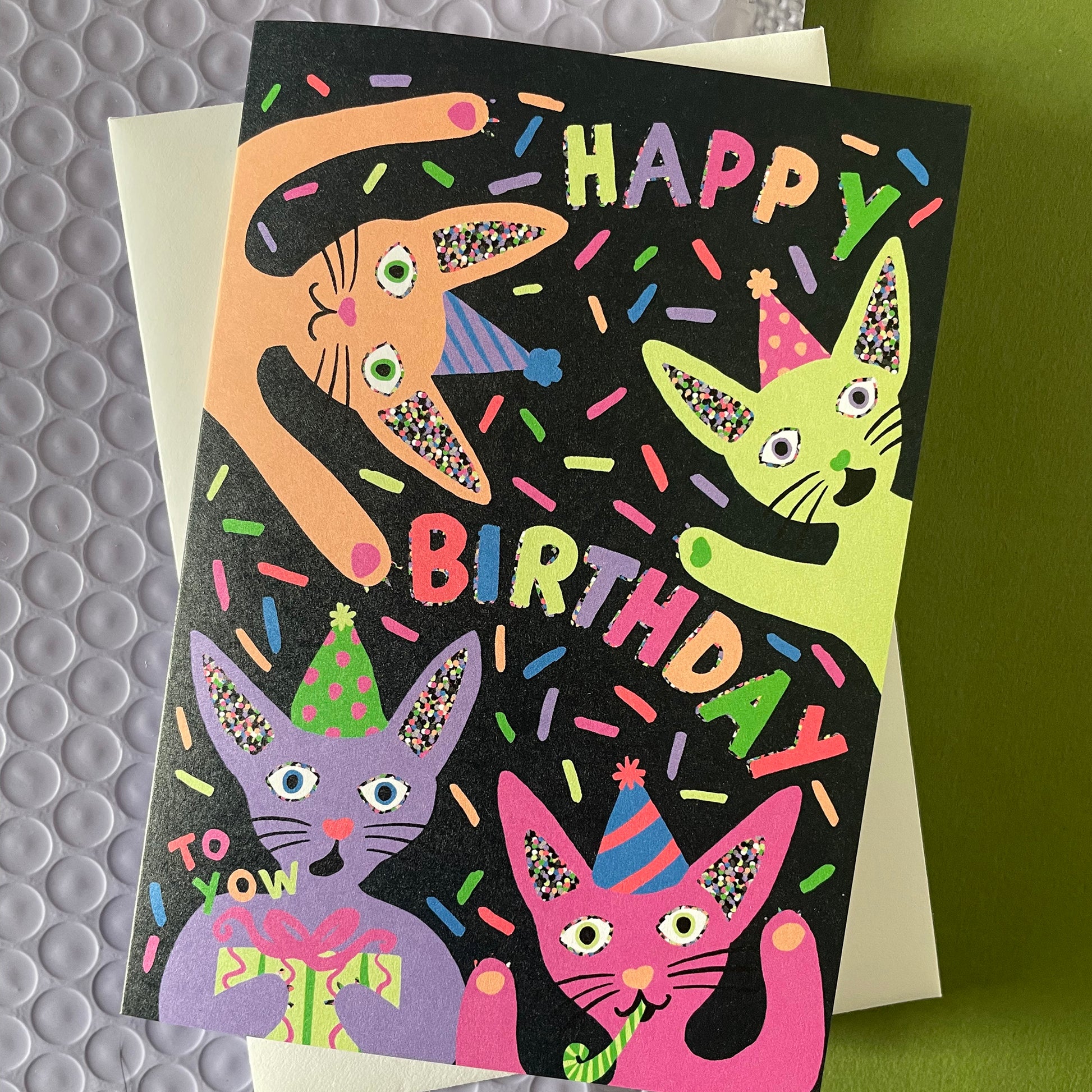Image shows a quirky black greetings card with neon, illustrated cats in party hats and the words happy birthday.