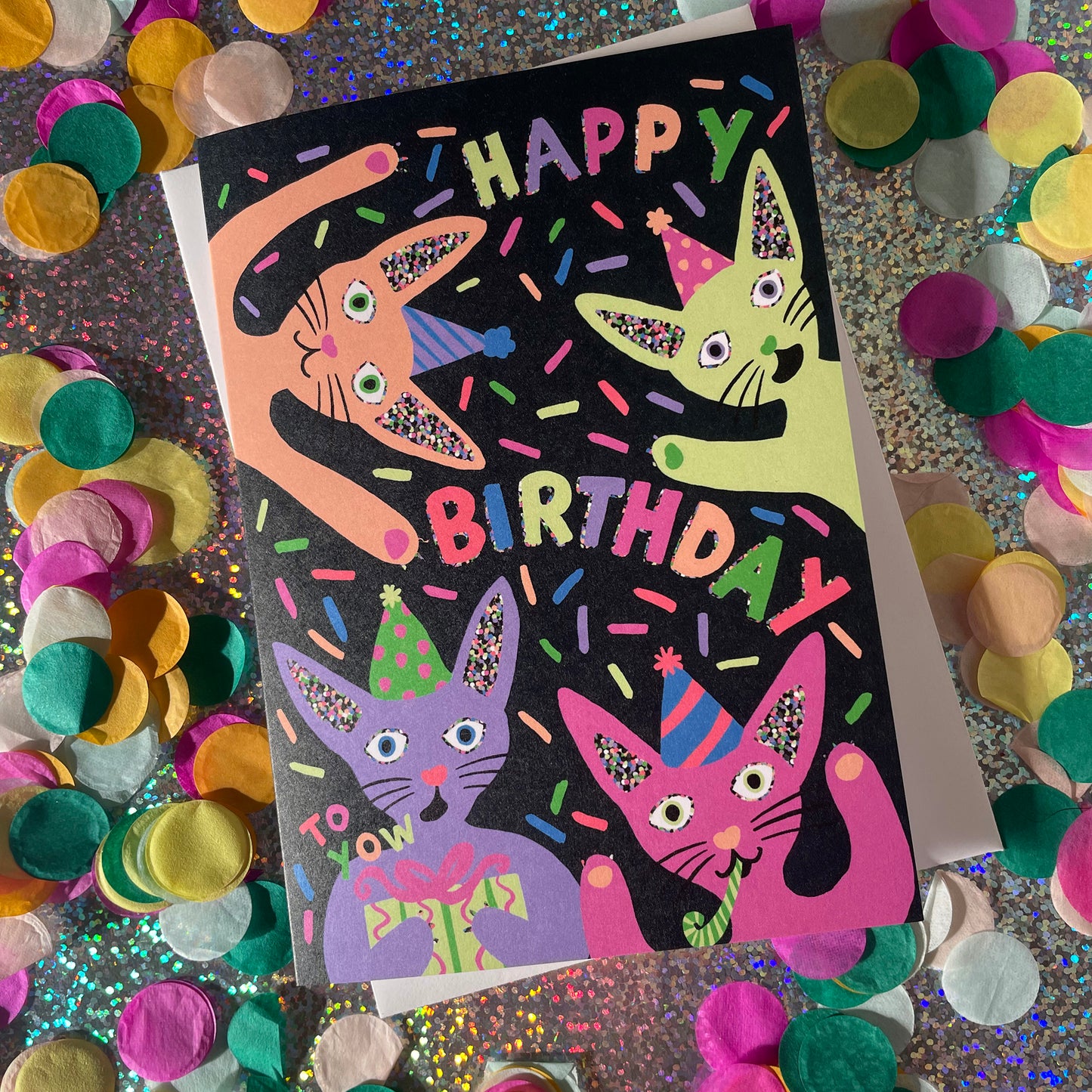 Image shows a quirky black greetings card with neon, illustrated cats in party hats and the words happy birthday.