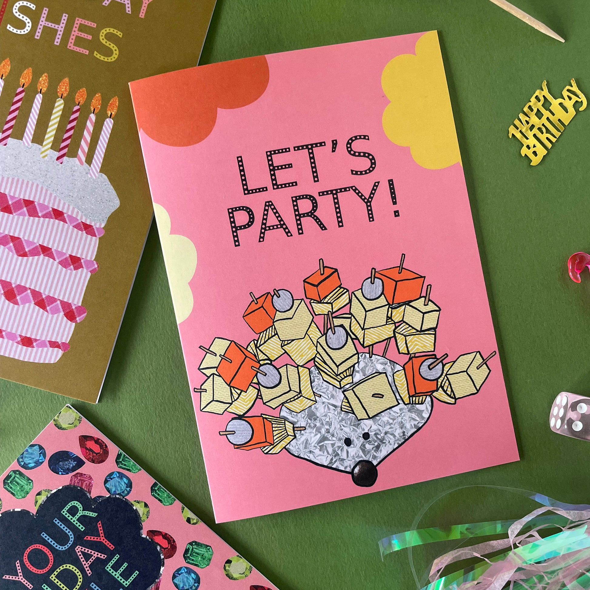 Image shows a pink greetings card with an illustration of a silver foil 'party hedgehog' with cheese, pineapple and pickled onions on cocktail sticks and the words 'let's party'