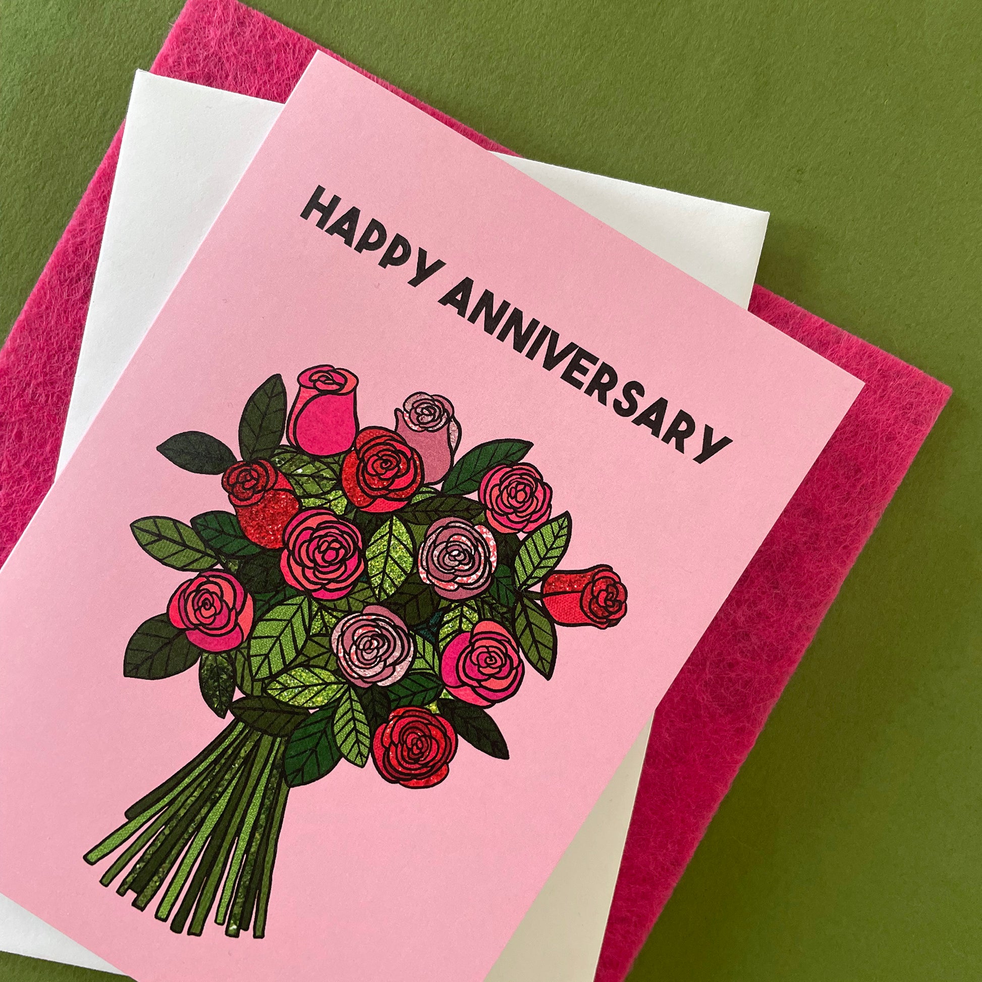 Image shows a light pink greetings card with an illustration of a bunch of roses and the words 'Happy Anniversary'